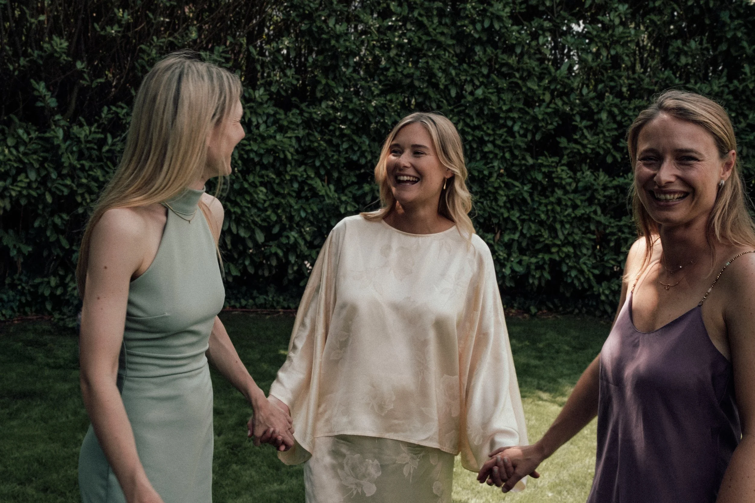 Three women holding hands and smiling outdoors, with a lush green hedge in the background.