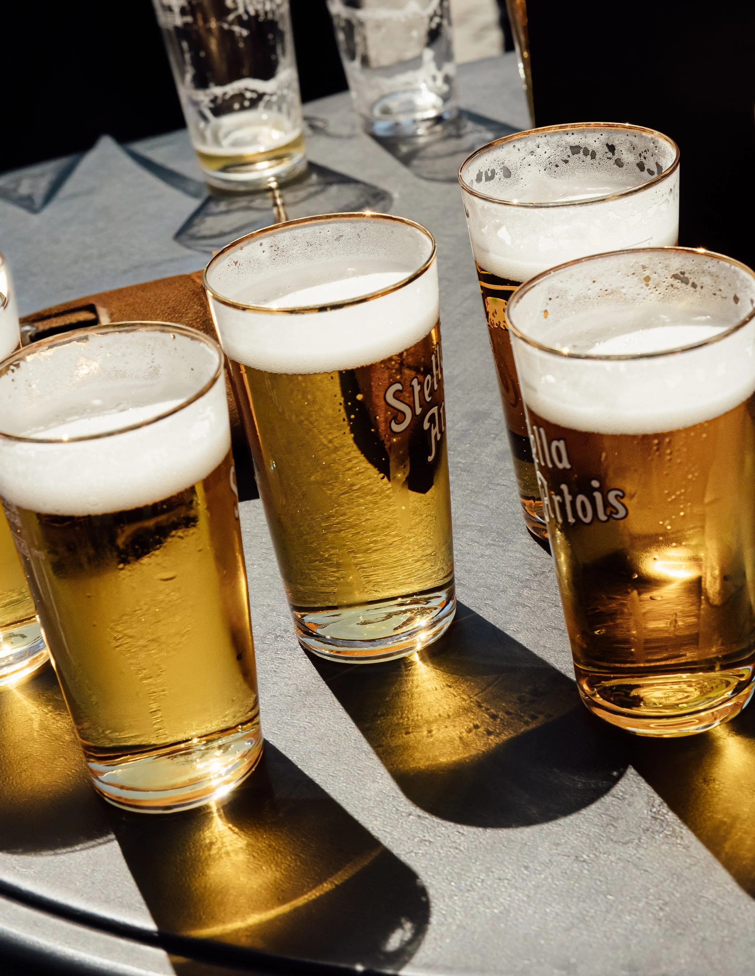 Multiple glasses of beer on a dark table, some filled with foam, casting shadows on the surface.