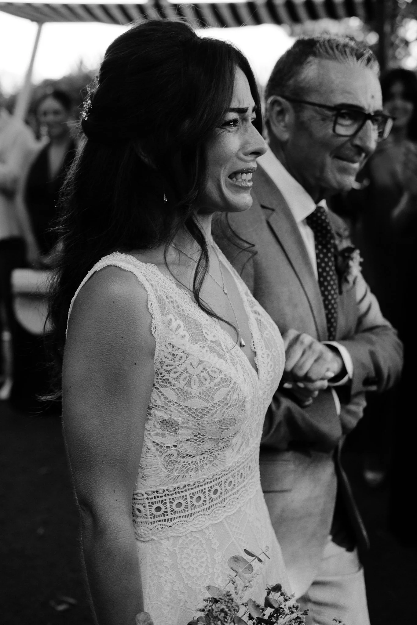 A woman in a white lace dress smiling and looking to the side, standing next to a man in glasses and a suit, with other people in the background at an outdoor event.