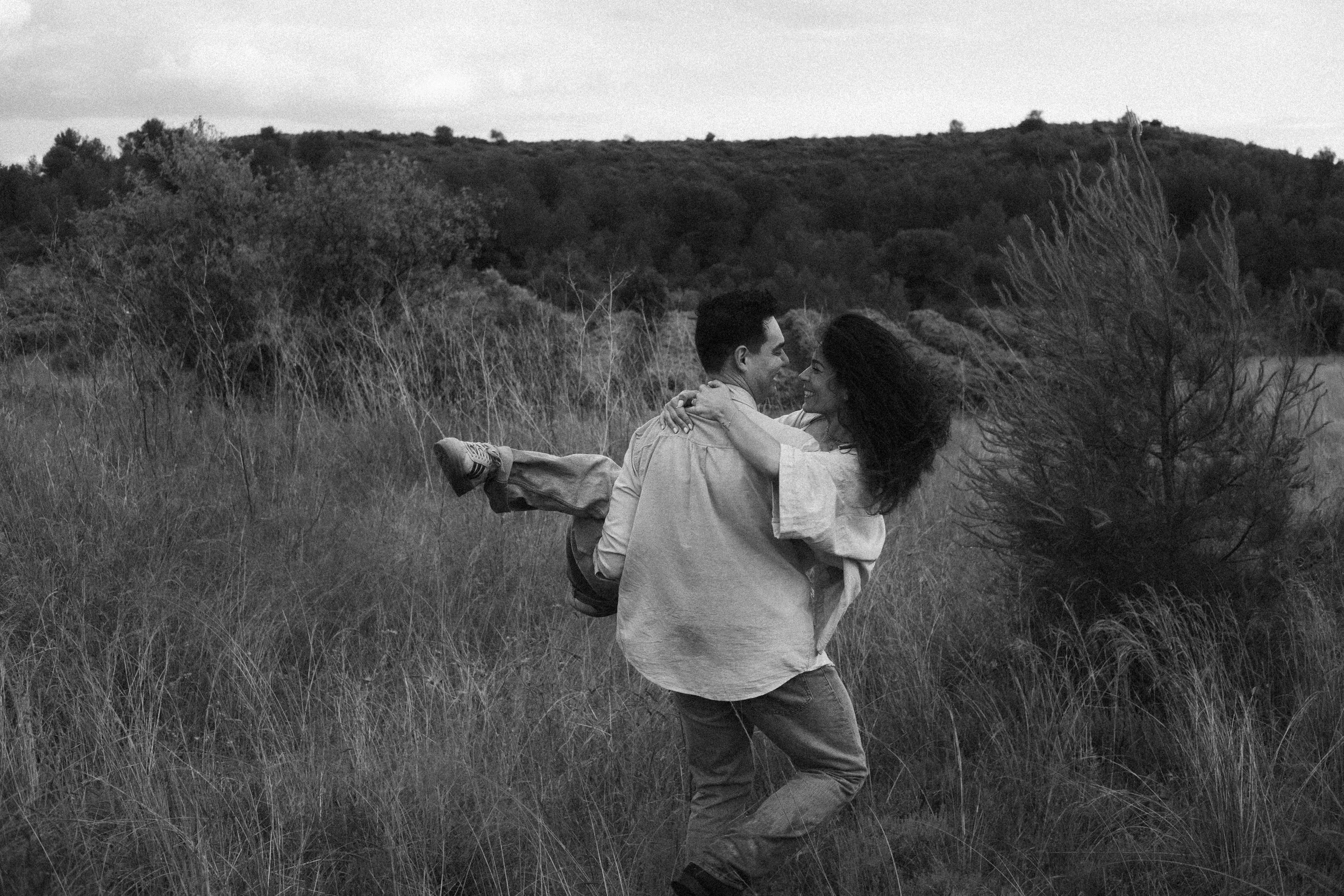 A couple is outdoors in a grassy field, with the man lifting the woman in his arms. Both are smiling and looking at each other, with a backdrop of trees and hills under a cloudy sky.