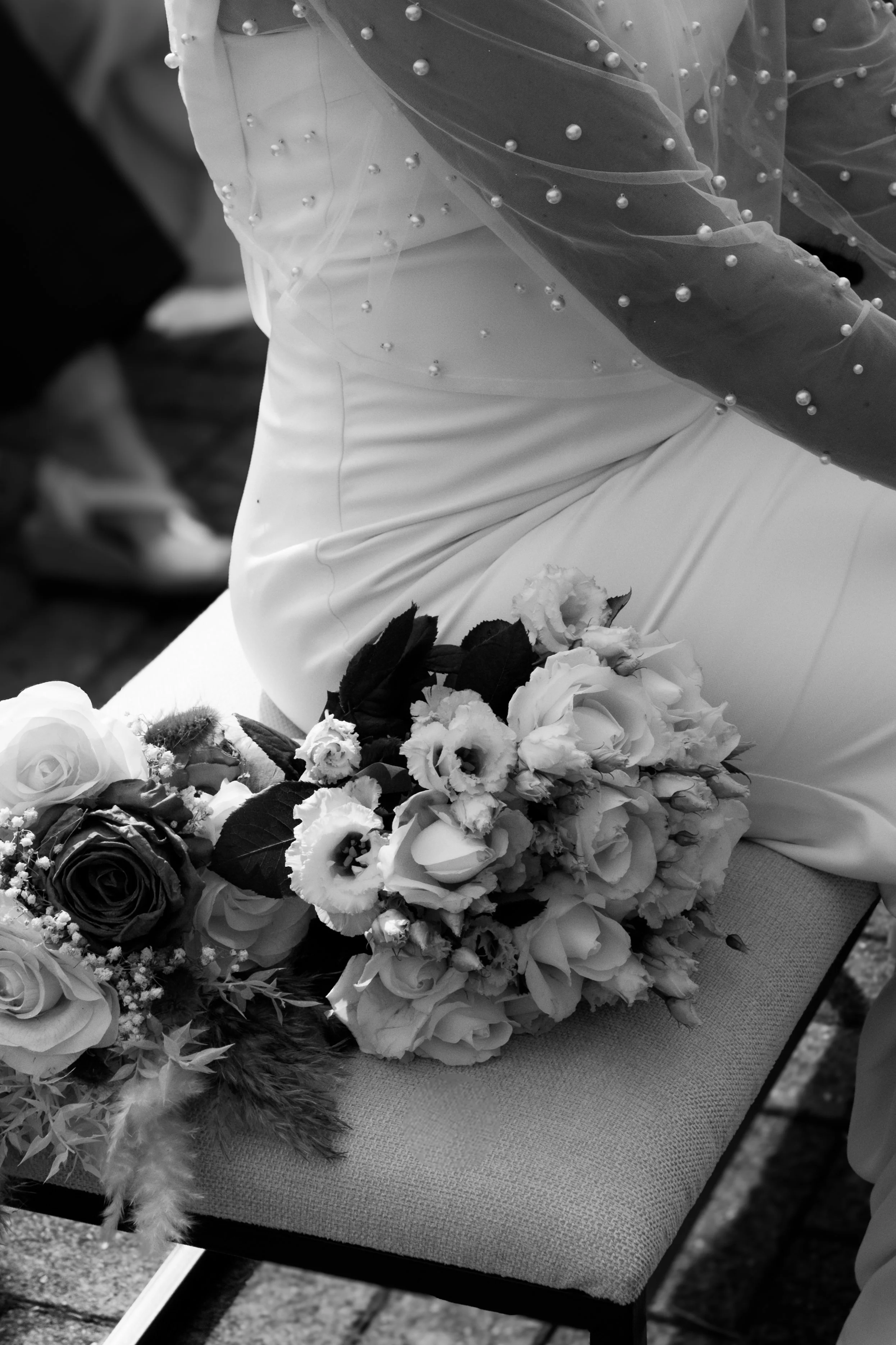 Close-up of a wedding bouquet with various flowers resting on a cushioned surface, with a bride and groom sitting nearby, blurred in the background, in black and white.