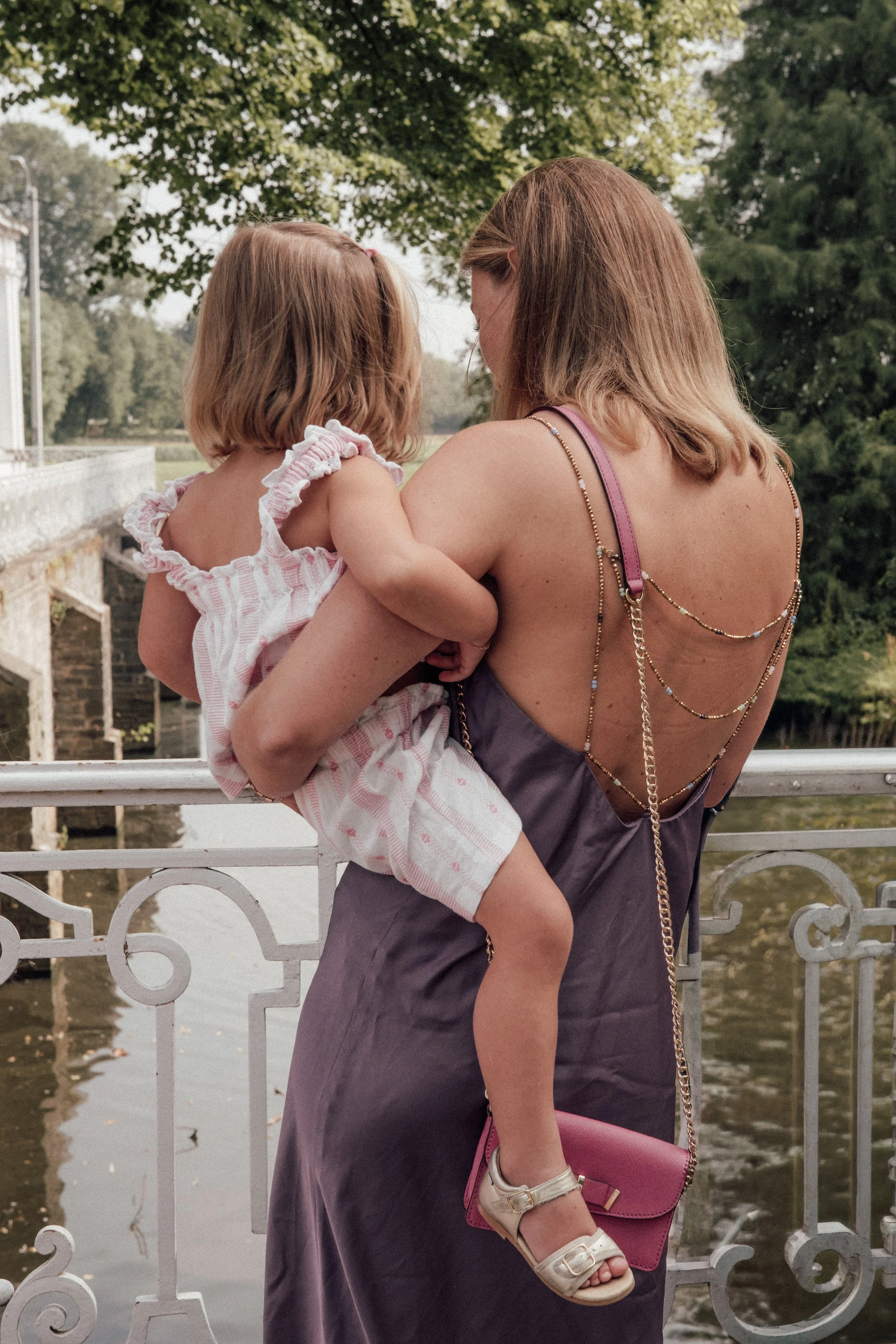 A woman holding a young girl over a river or pond with a decorative white metal railing, surrounded by green trees, on a cloudy day.