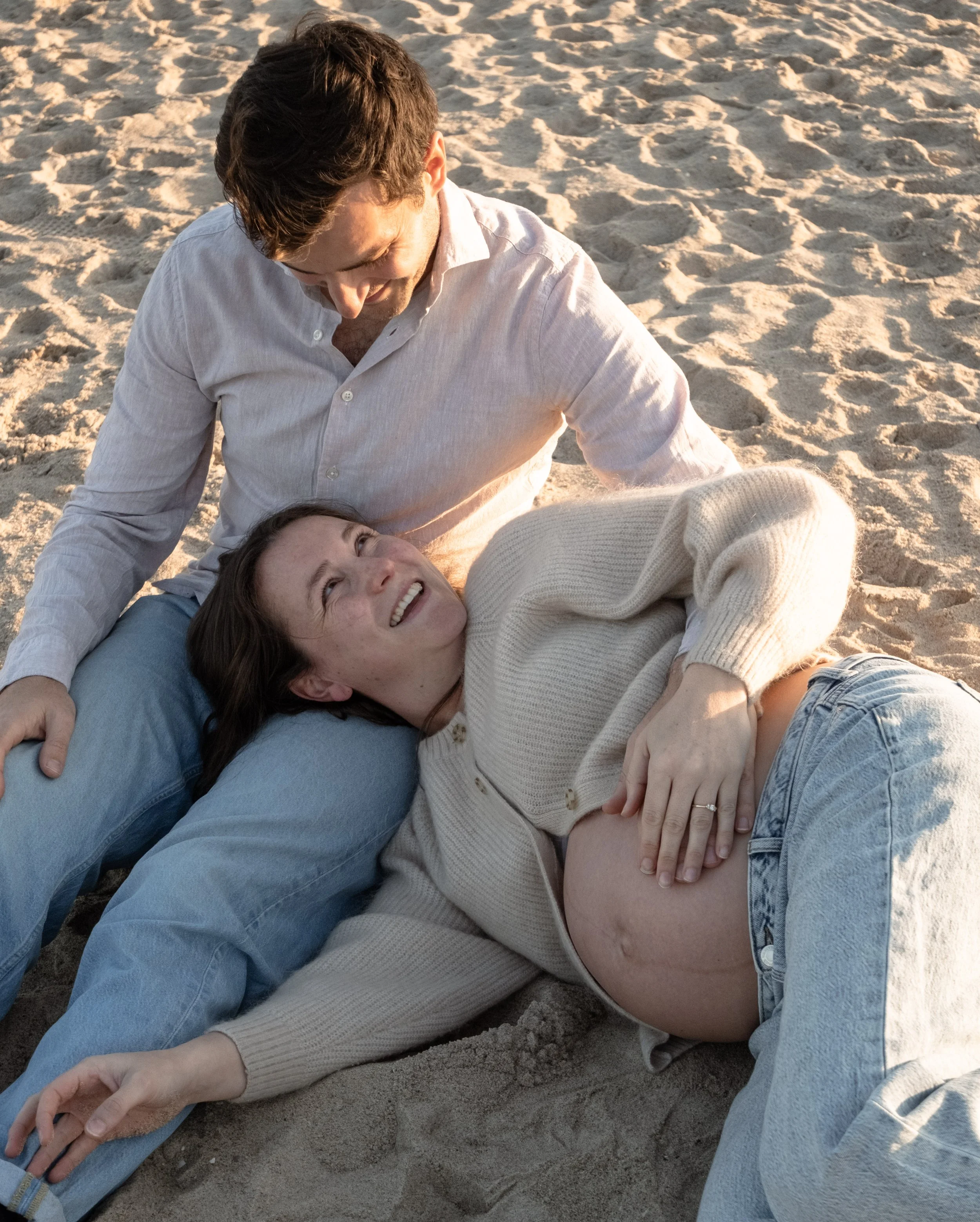 A smiling woman lying on the sand with her head resting on a man's lap. The man is sitting and wearing a white shirt, looking down at the woman with a joyful expression. The scene is illuminated by warm sunlight, possibly during sunset, on a beach.