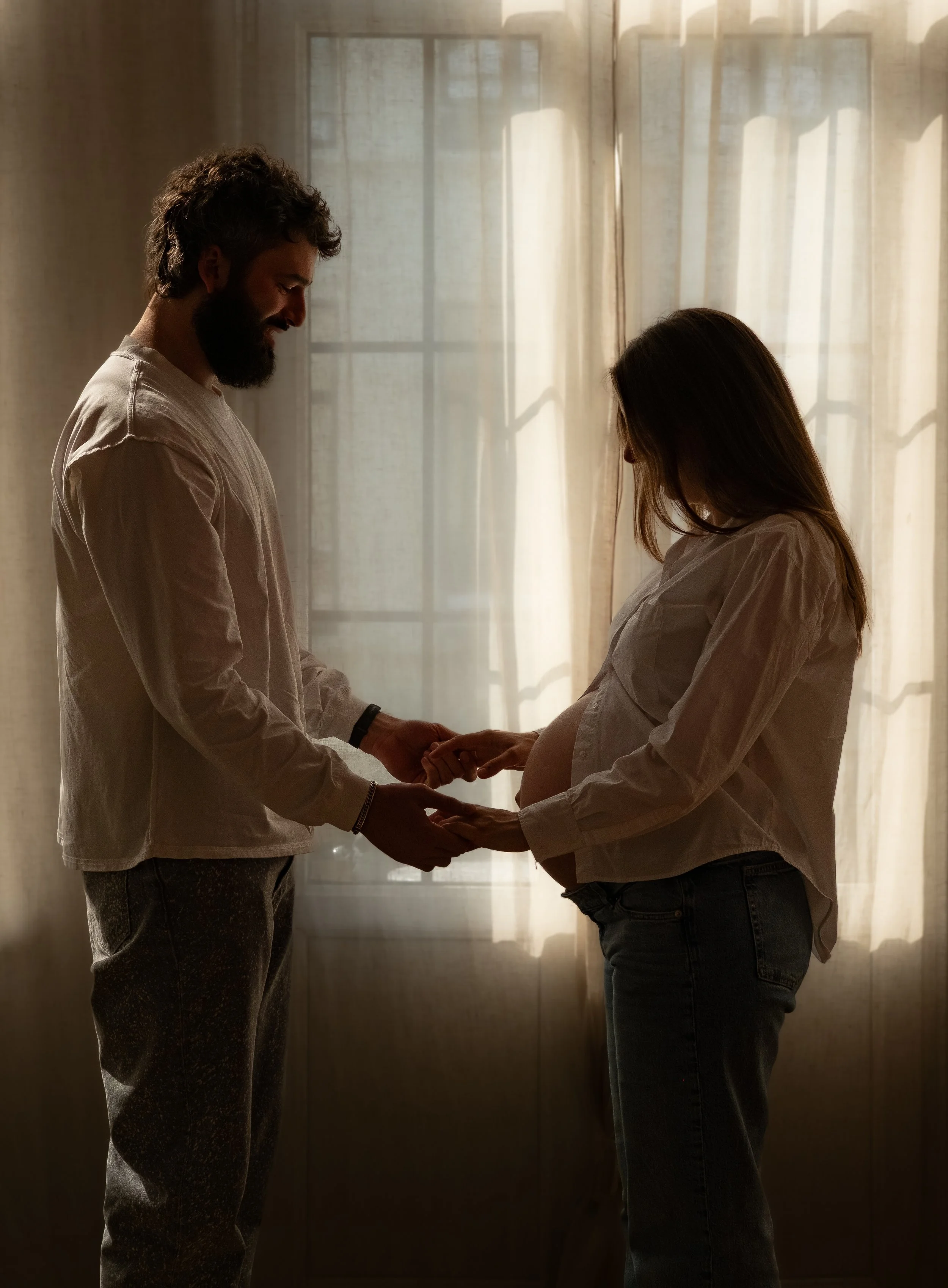 A man and a woman standing in front of a window with curtains, holding hands and smiling at each other, with a pregnant woman showing her baby bump.