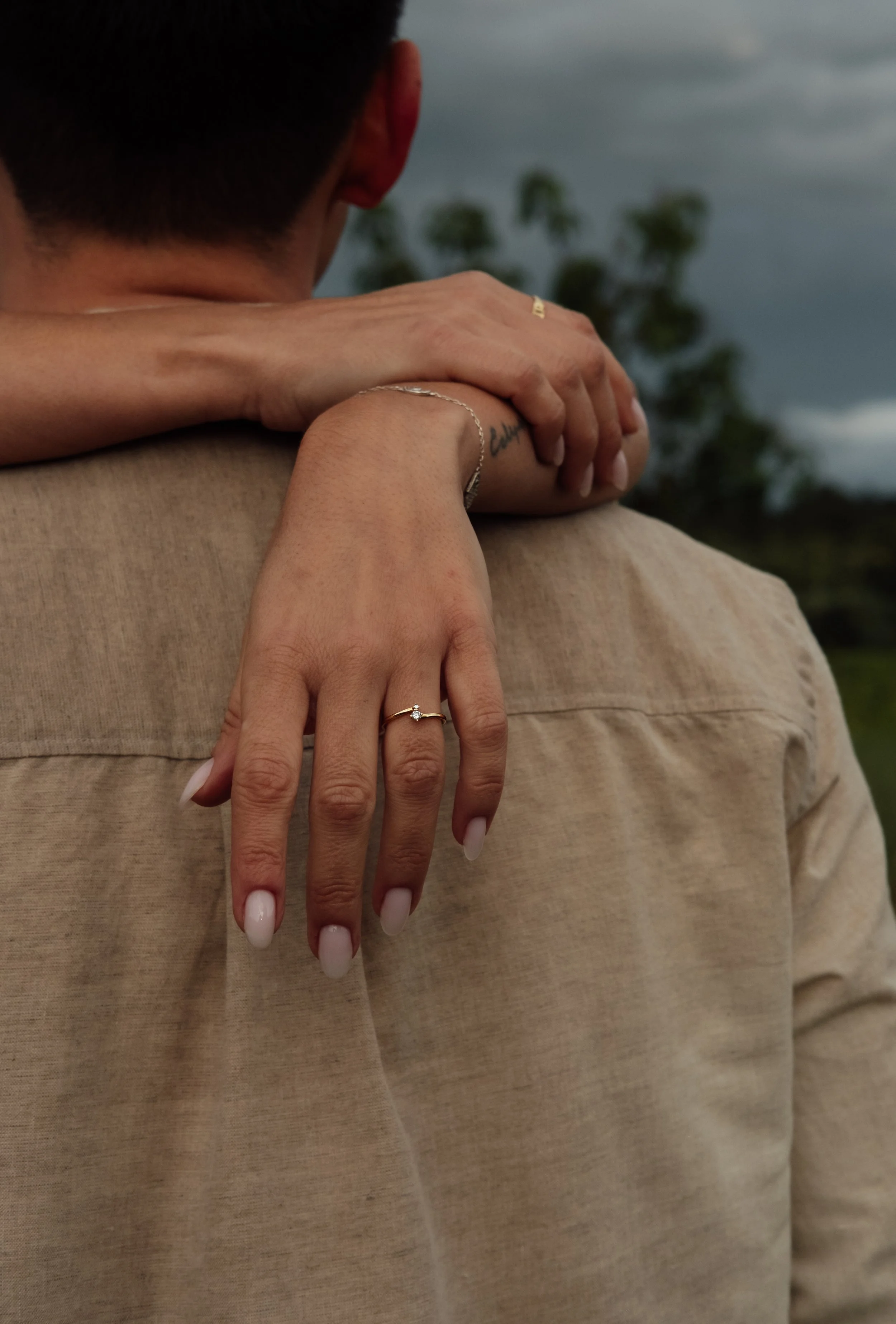 A close-up of a couple embracing, with the woman's hand resting on the man's shoulder, showing rings on her fingers. The background includes trees and a cloudy sky.