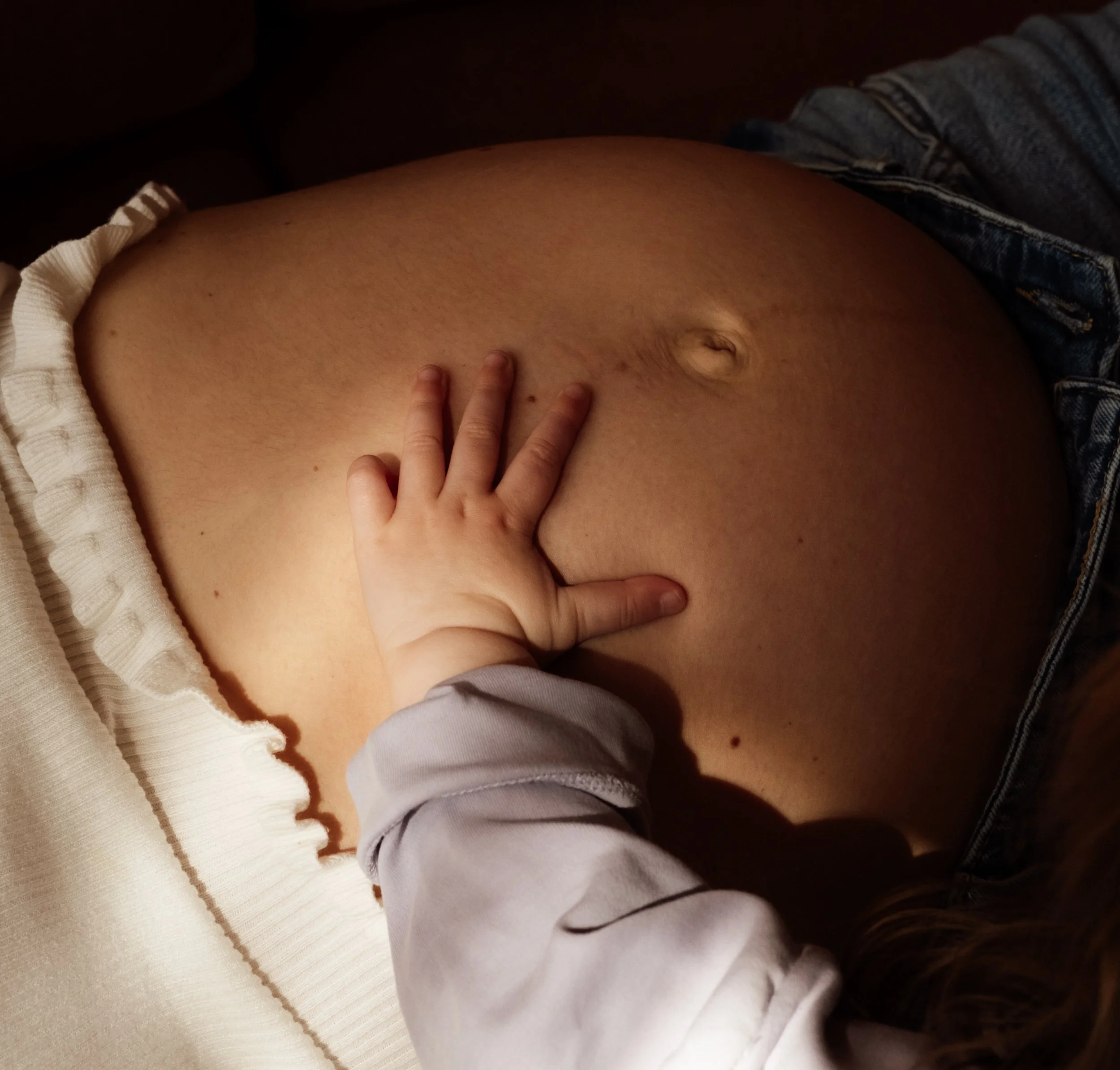 Close-up of a person's pregnant belly with a hand resting on it, wearing casual clothing including a white shirt and denim shorts.