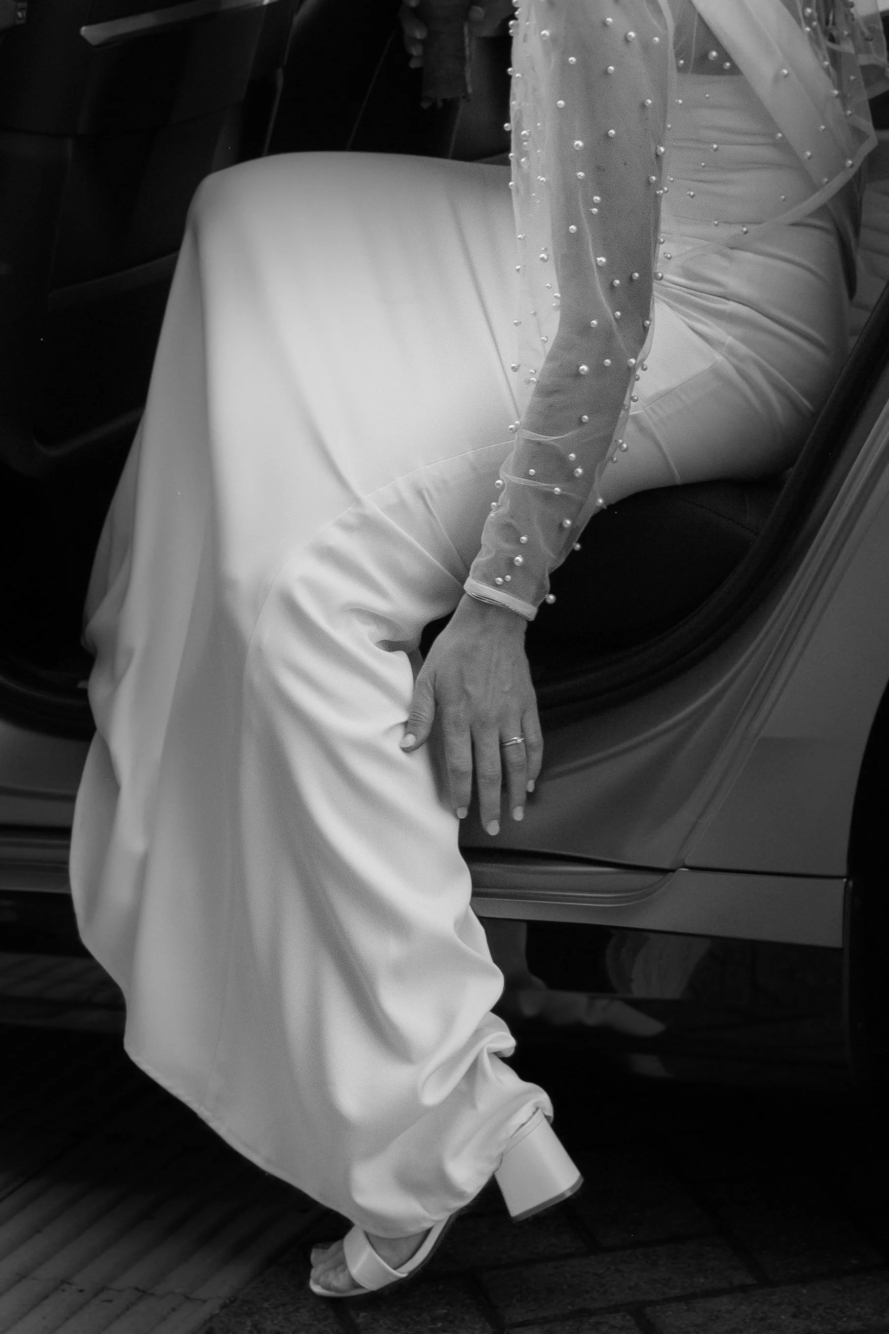A bride wearing a white dress with pearl embellishments sitting in the backseat of a car, with their hand resting on the door frame and wearing high-heeled shoes.