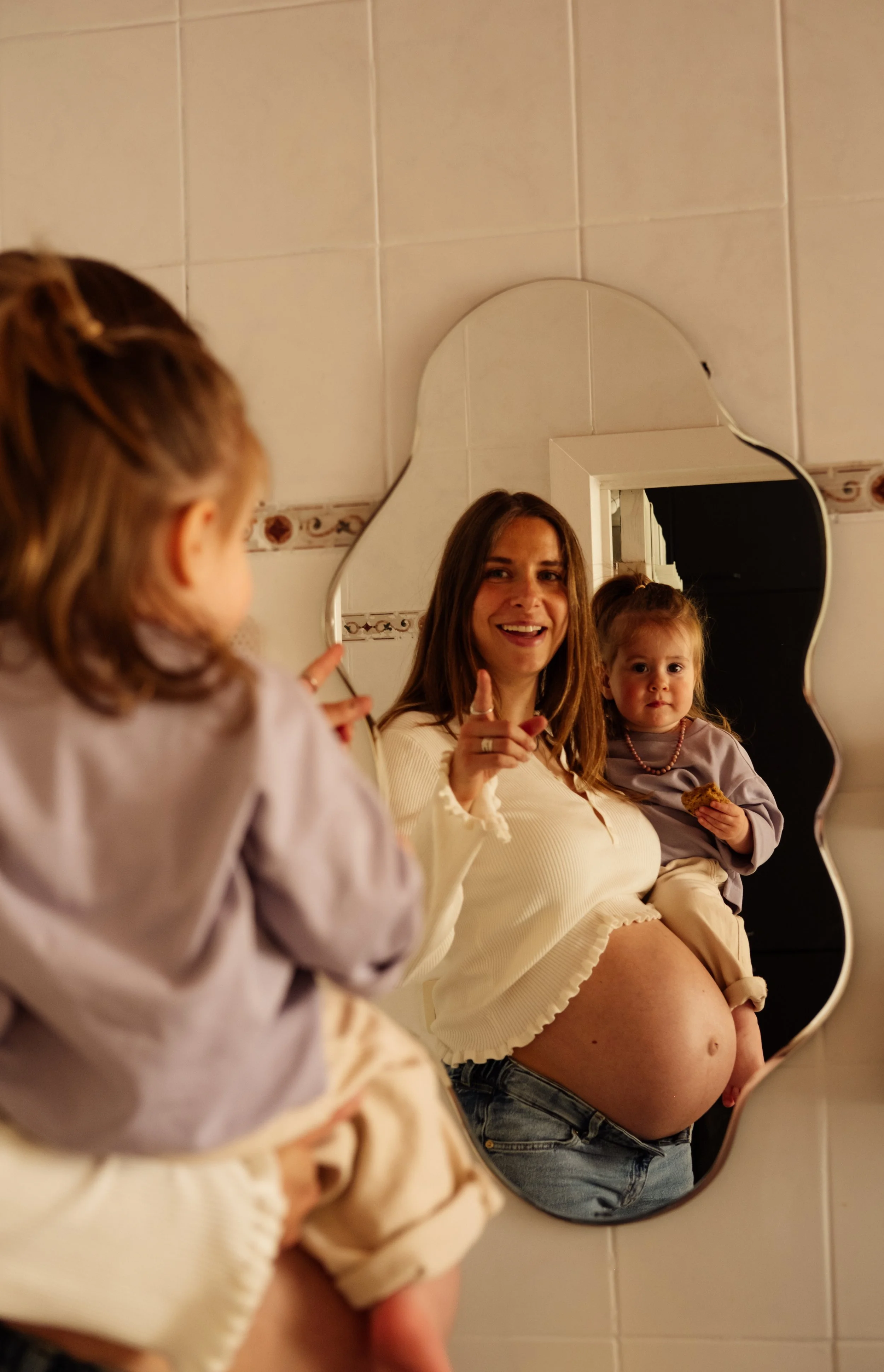 A woman and two young girls sitting in front of a mirror in a tiled bathroom, with the woman showing her pregnant belly and smiling.