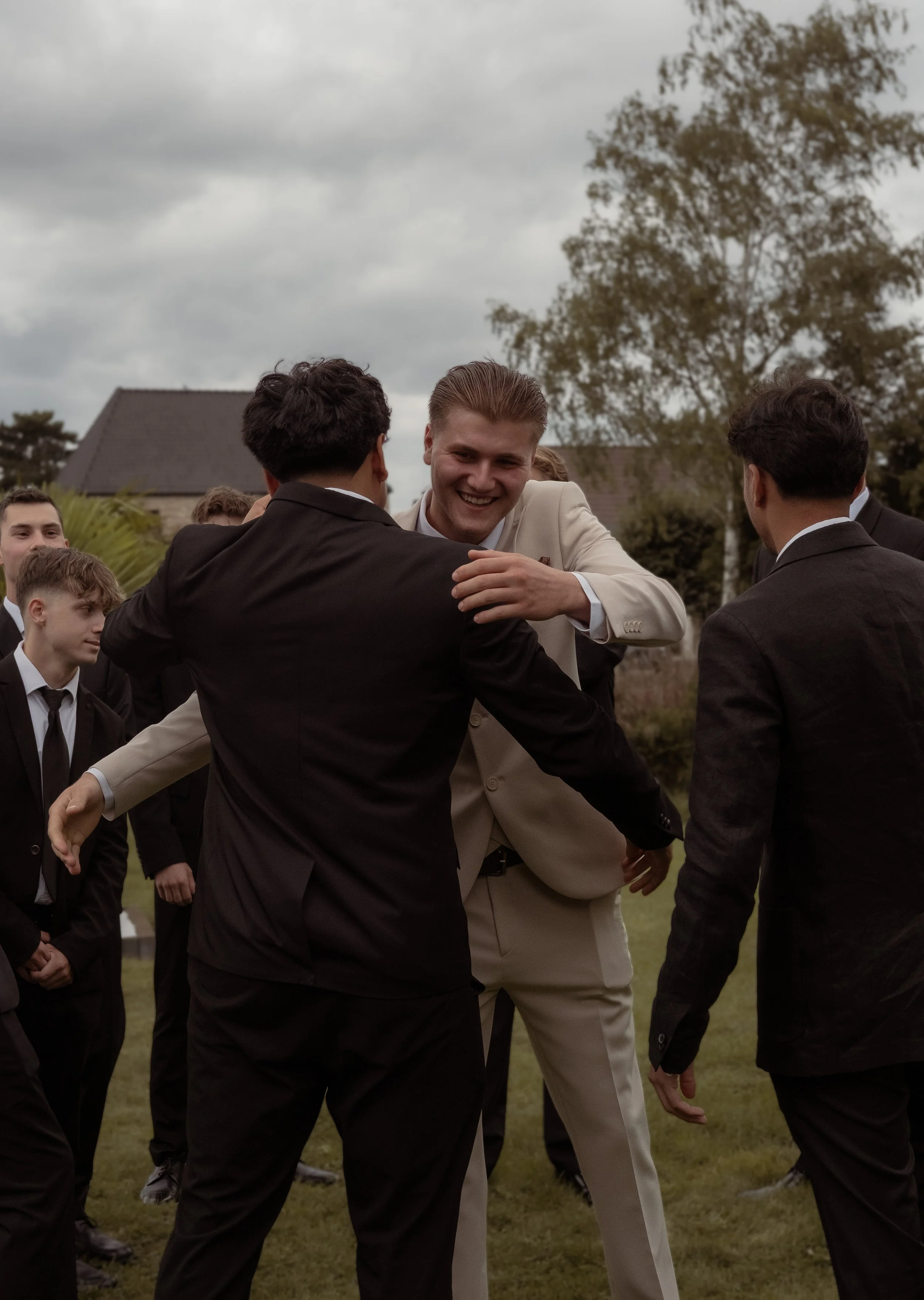 A group of men in suits are standing outdoors on a cloudy day, with one man in a white suit smiling and embracing another man in a dark suit.