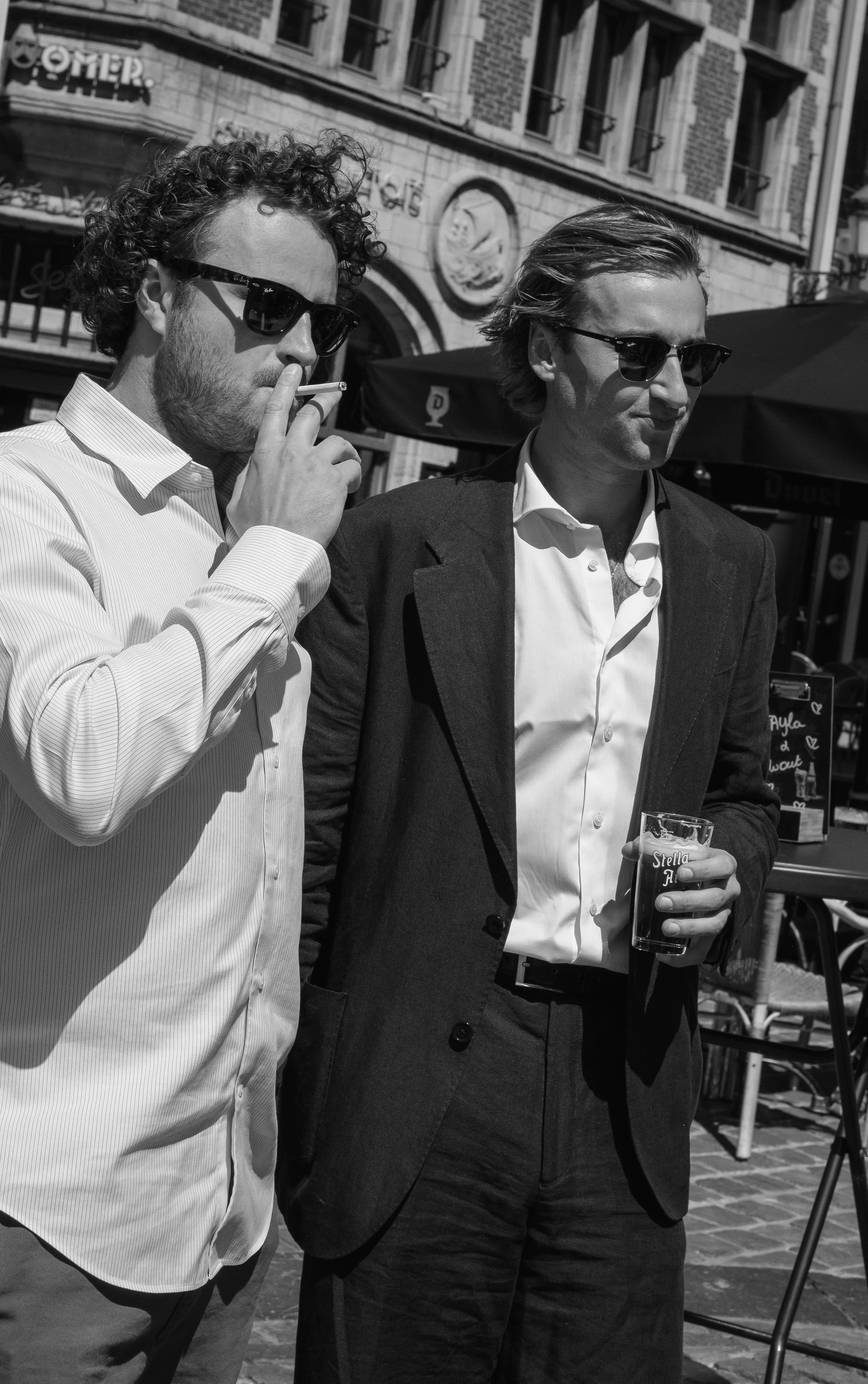 Two men in suits and sunglasses standing outdoors on a city street. One man is smoking a cigarette, and the other is holding a glass of beer. There are buildings and outdoor furniture in the background.