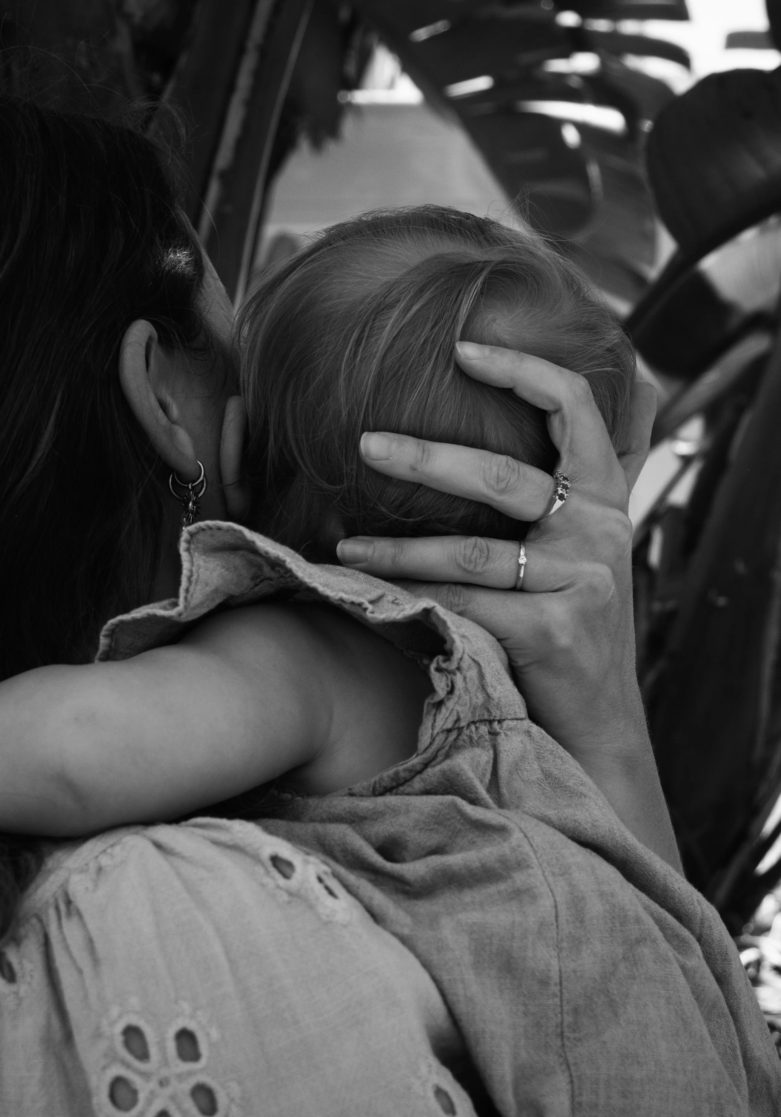 A woman embraces a child, holding the child's head tenderly with one hand. The woman has earrings and a ring, and her hair is styled back. The child has light-colored hair and wears a sleeveless top. The background shows folding chairs, suggesting an