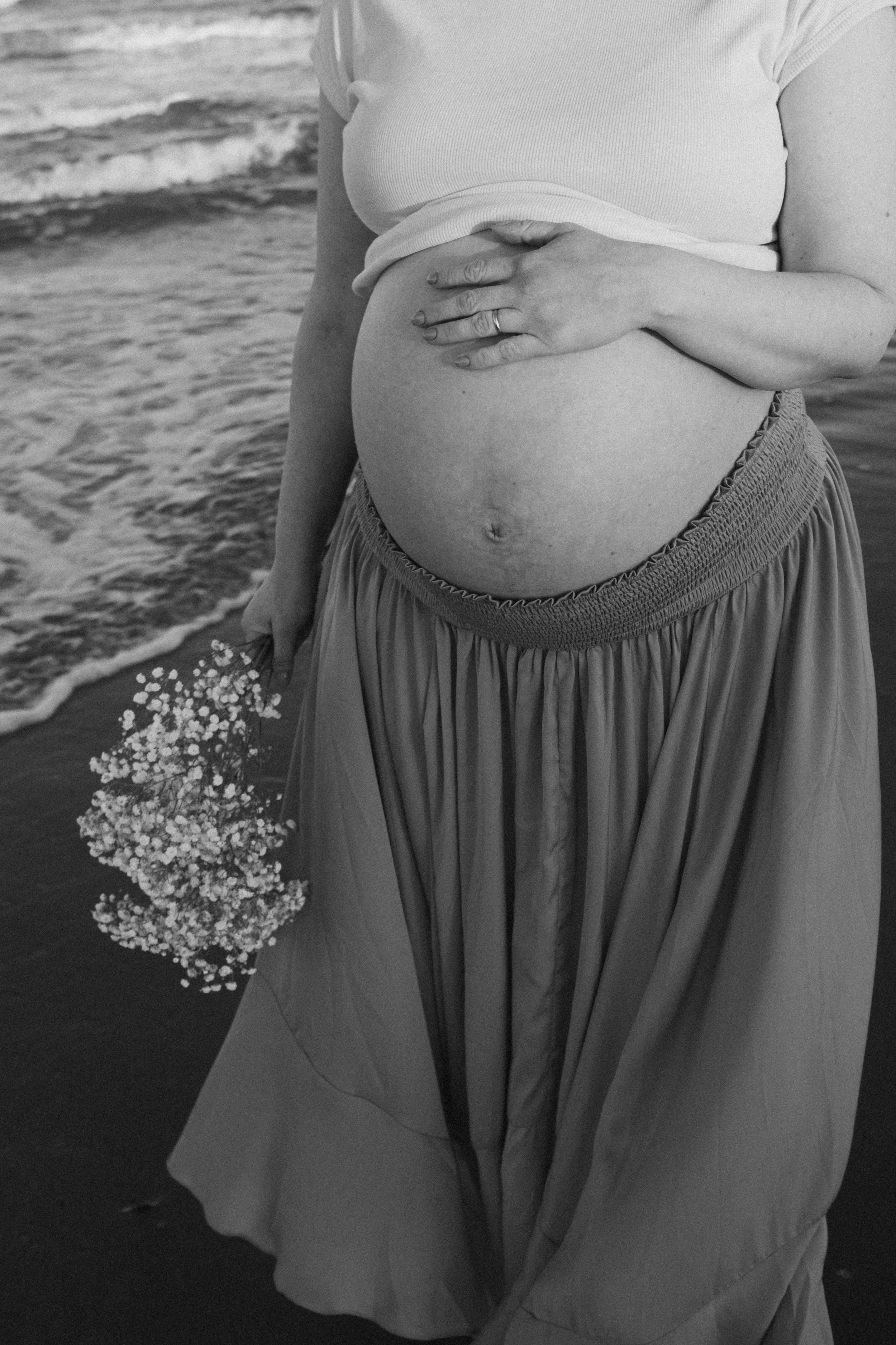 Black-and-white close-up of a pregnant woman holding her belly in a flowing dress beside a window.