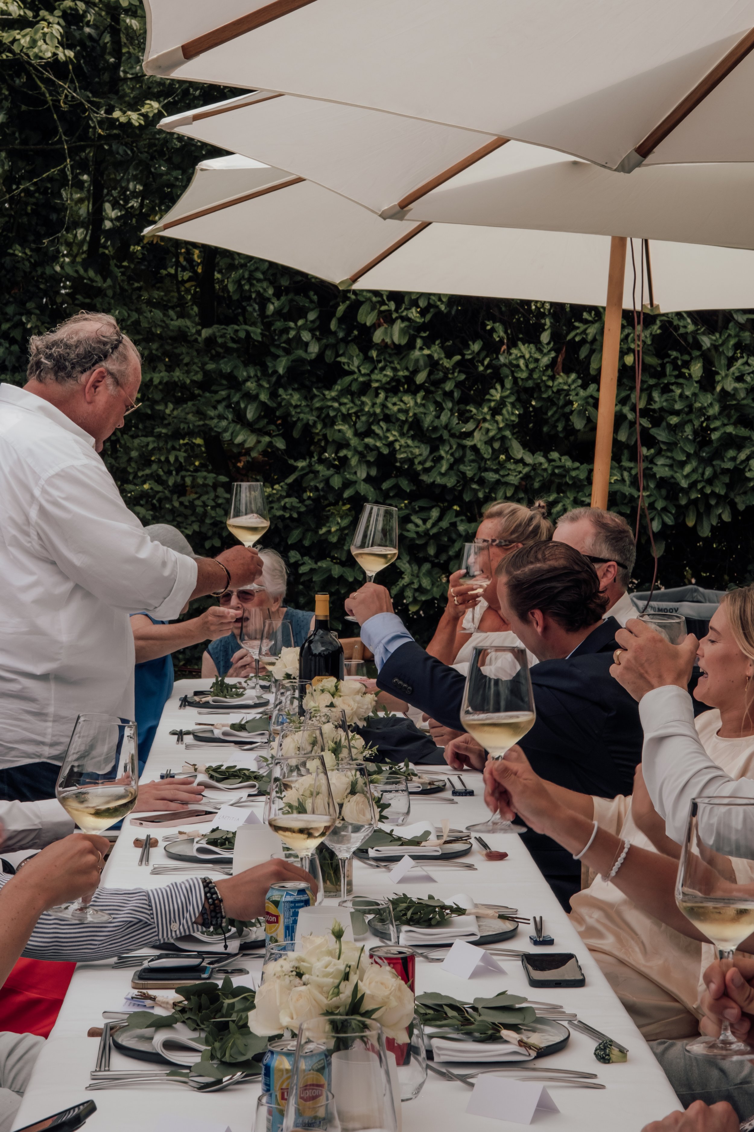 People seated at a long dining table outdoors, raising glasses in a toast, with white umbrellas overhead and greenery in the background.
