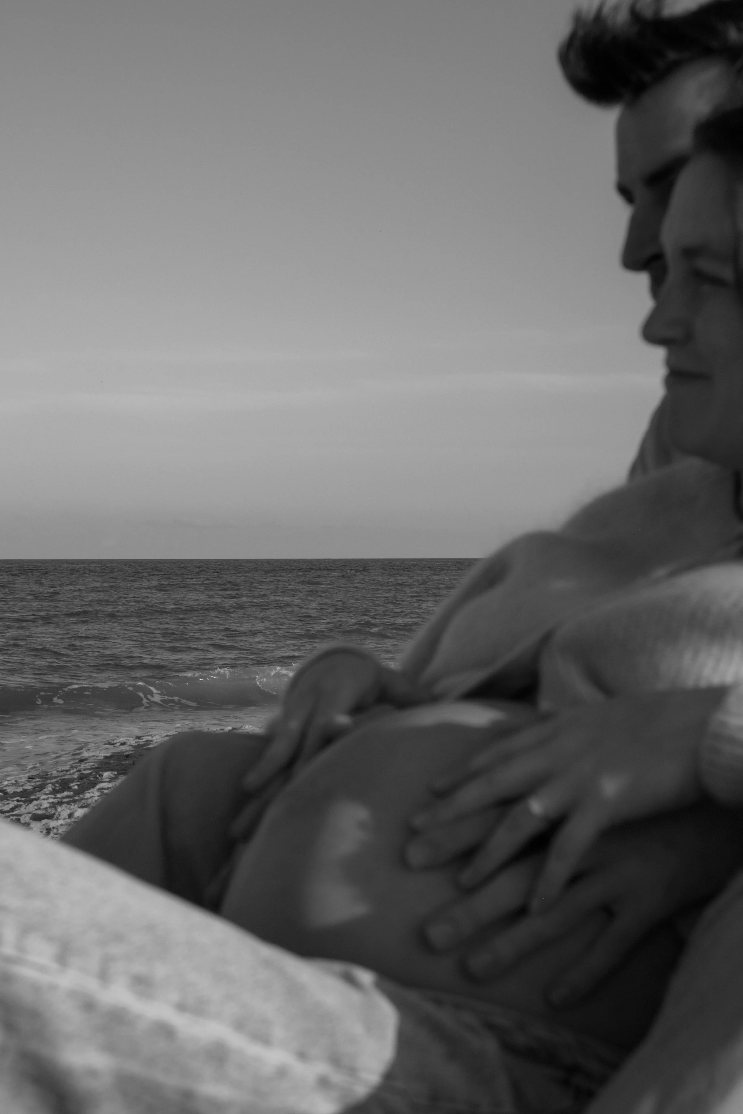 A black and white photo of a person wearing a hoodie, sitting by the ocean, looking into the distance.