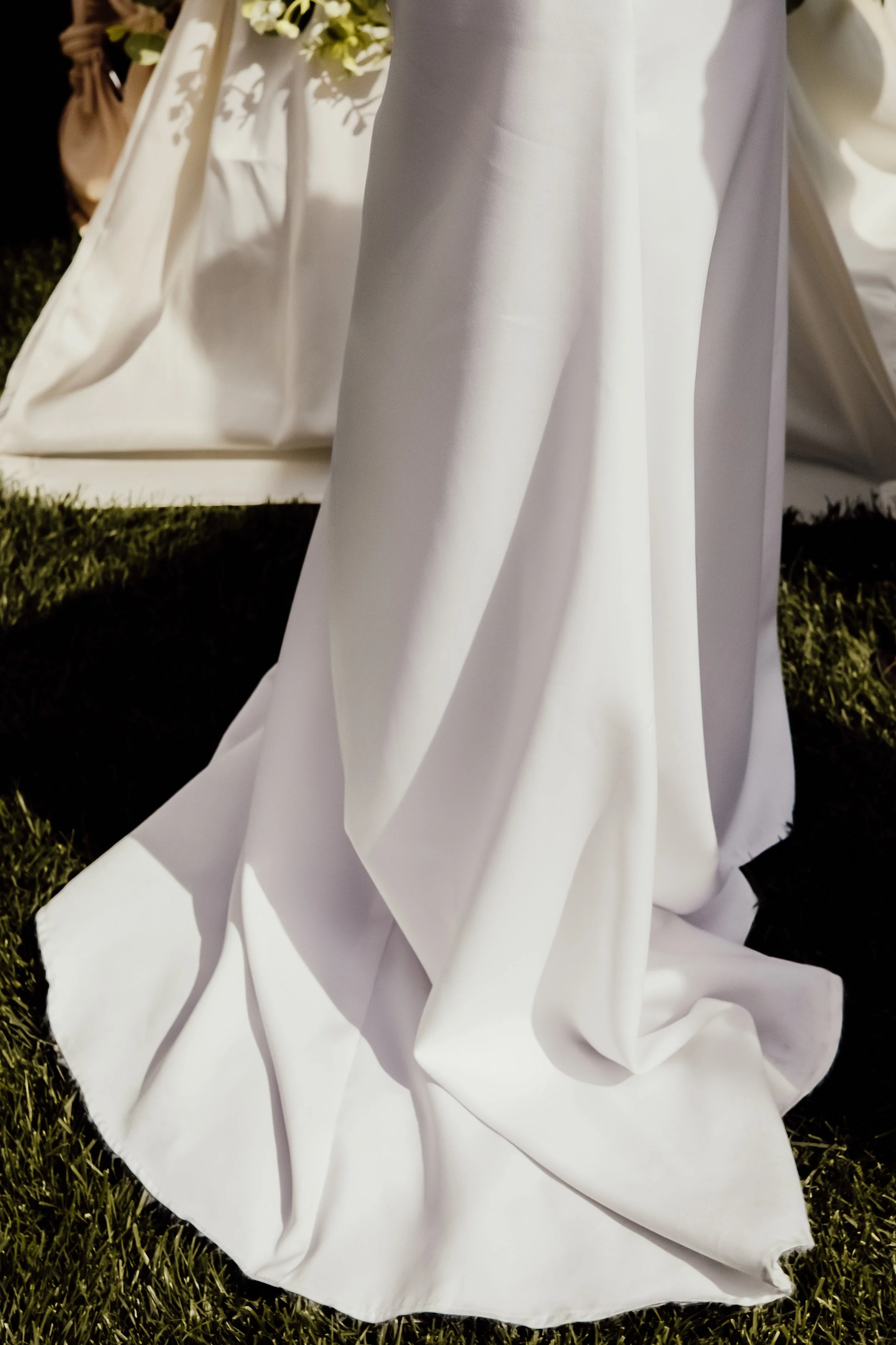 A white wedding dress with long sleeves and a full skirt is draped on the grass outdoors, with a background of white flowers and greenery.