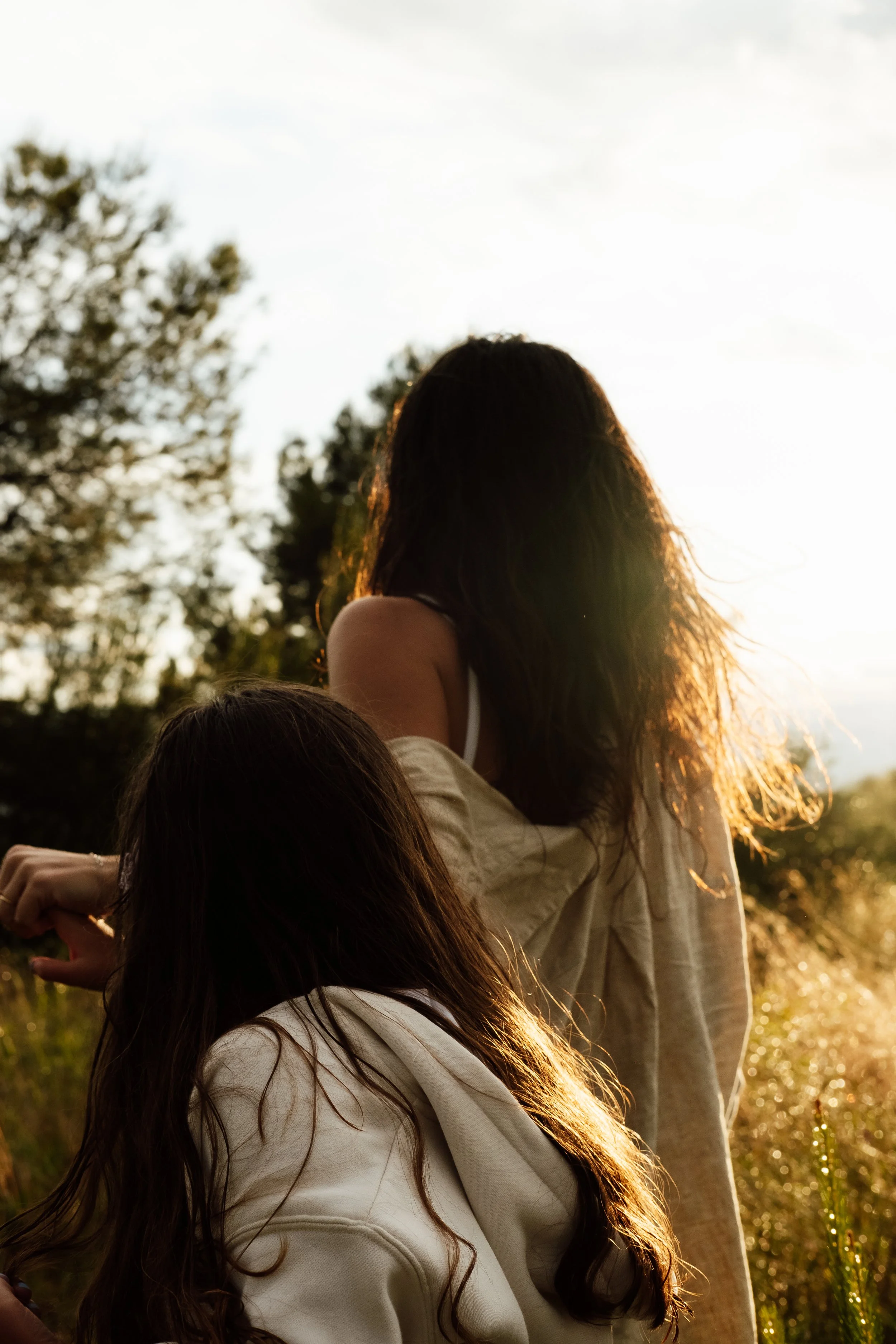 Two women with long brown hair, wearing light-colored clothing, are outdoors with trees in the background, illuminated by sunlight.