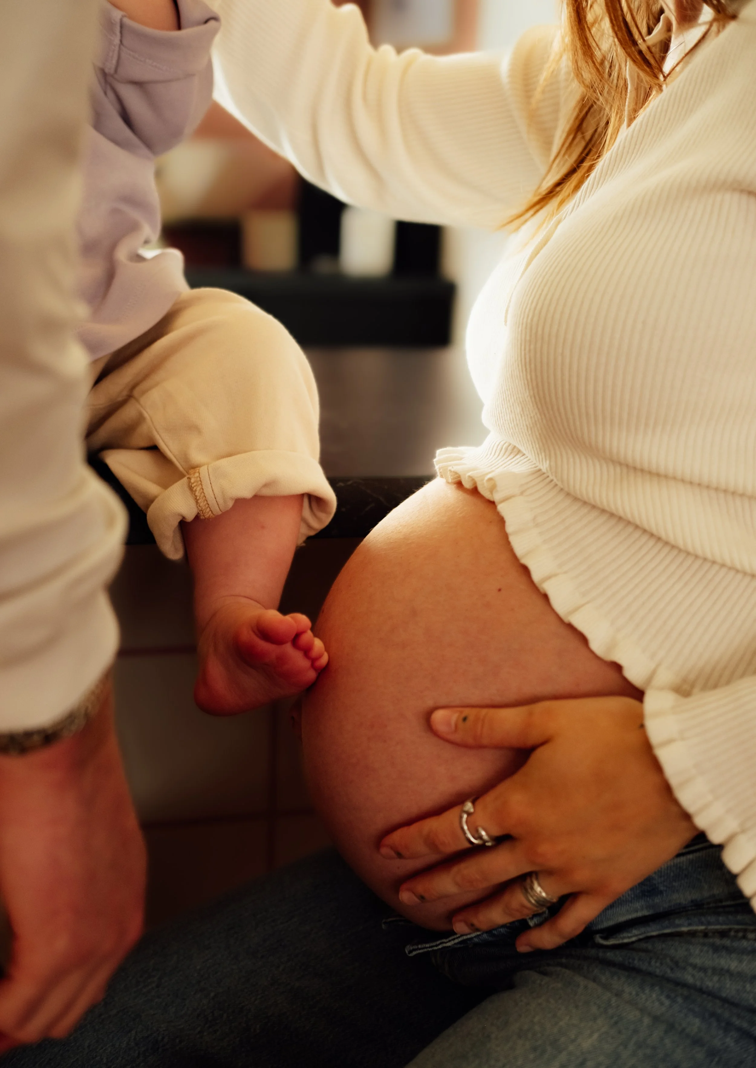Close-up of a pregnant woman and a young child, with the child touching the woman's pregnant belly. The woman is wearing a cream ribbed top, and the child is dressed in a beige shirt. The background is blurred.