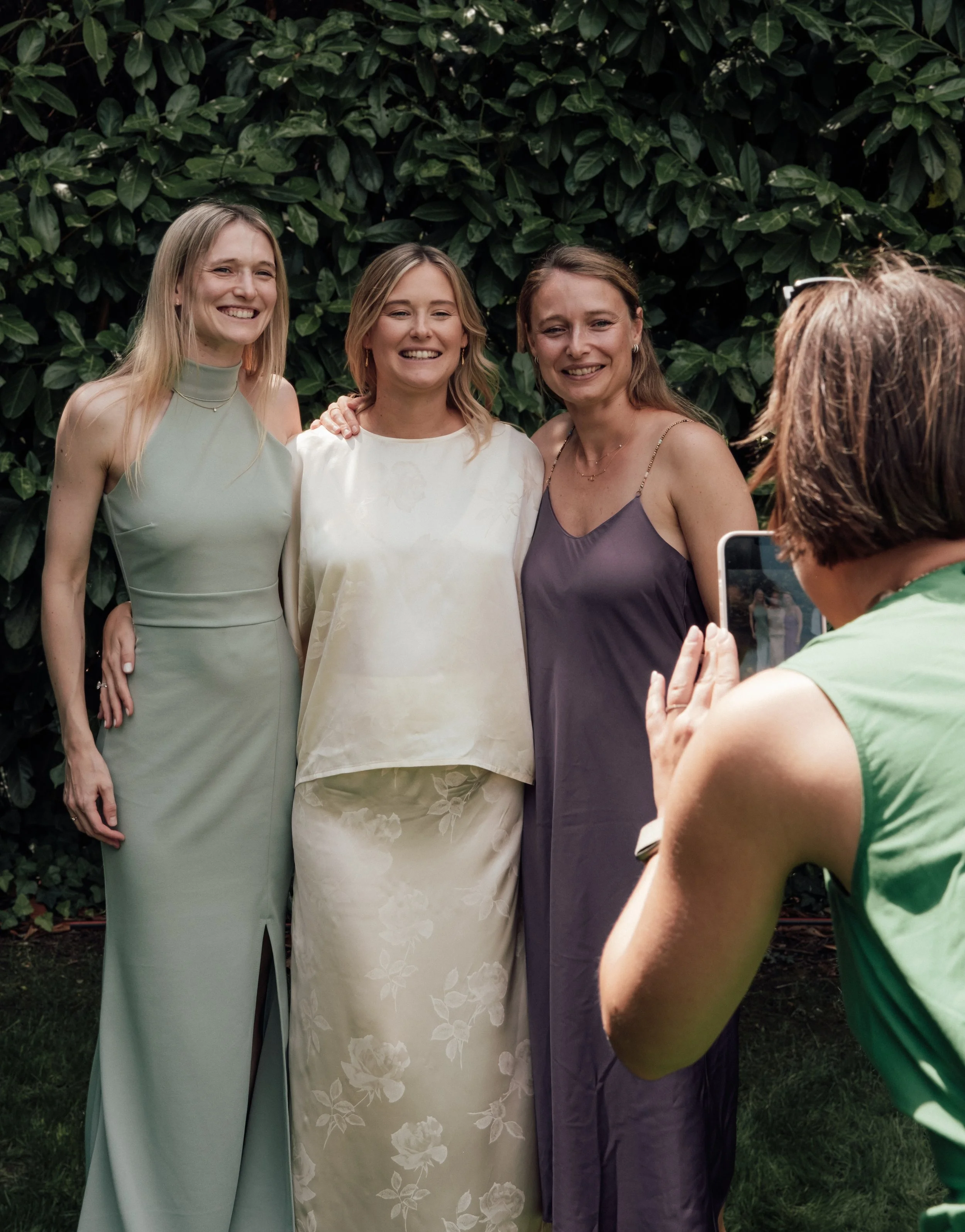 Three women in elegant dresses standing outdoors in front of a leafy green hedge, smiling for a photograph, with another woman in the foreground taking their picture with a smartphone.