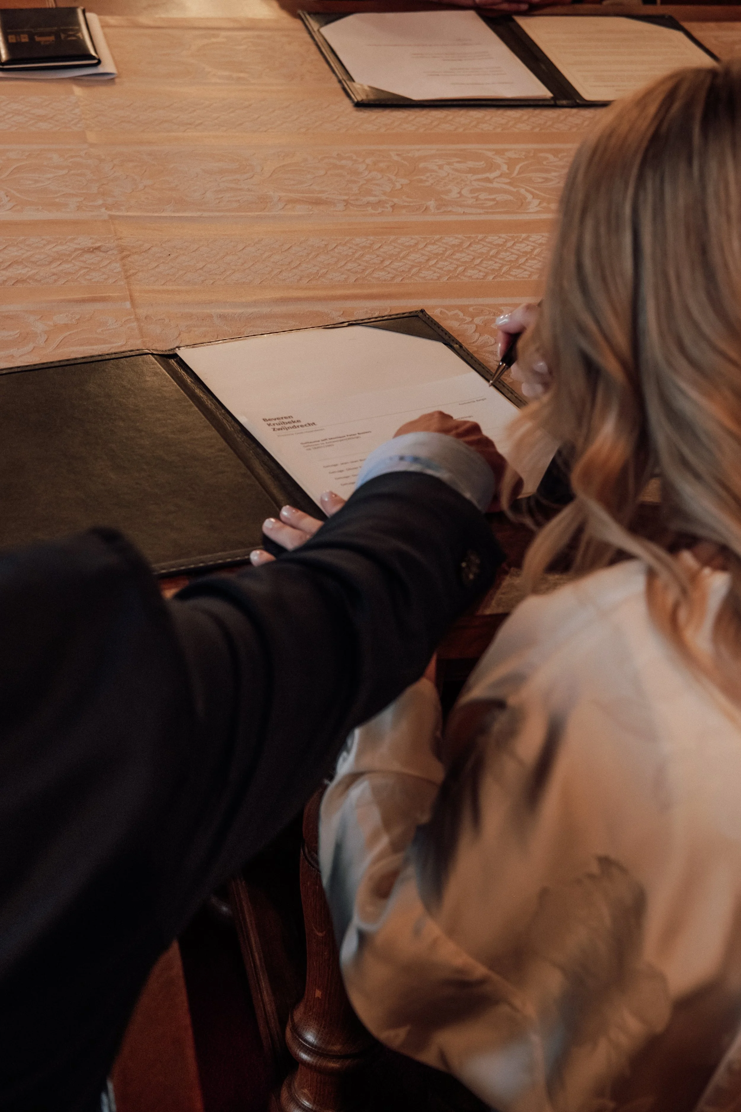 Person with long blonde hair writing on a document placed on a dark leather folder at a wooden table.