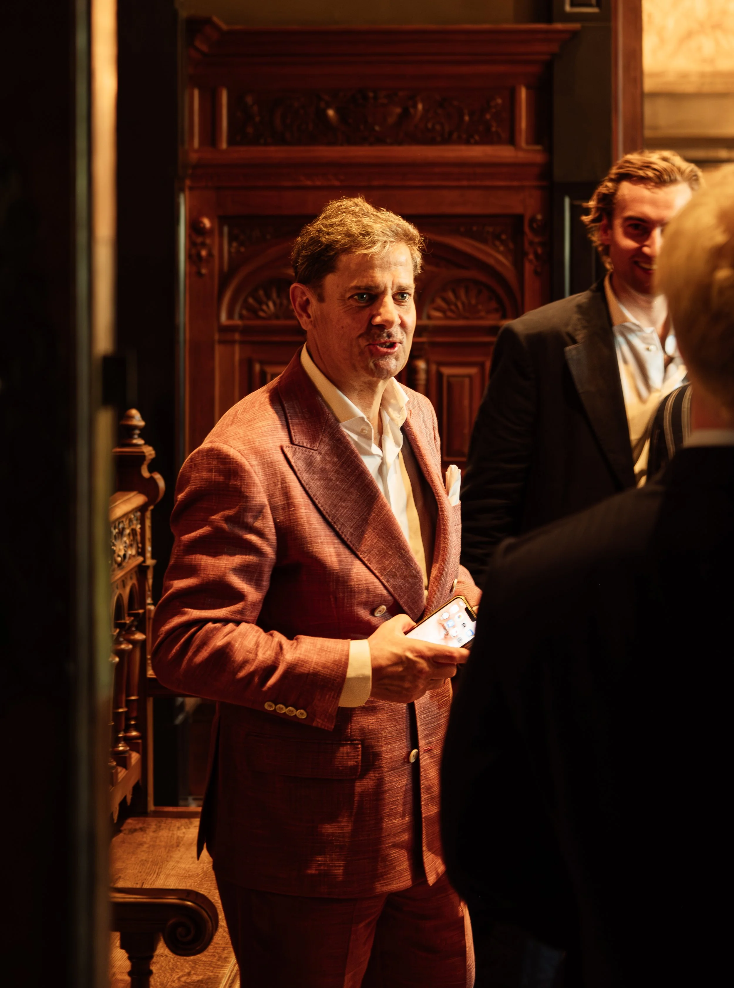 A man in a reddish-brown plaid suit holding a smartphone, standing in a dimly lit room with dark wooden paneling.