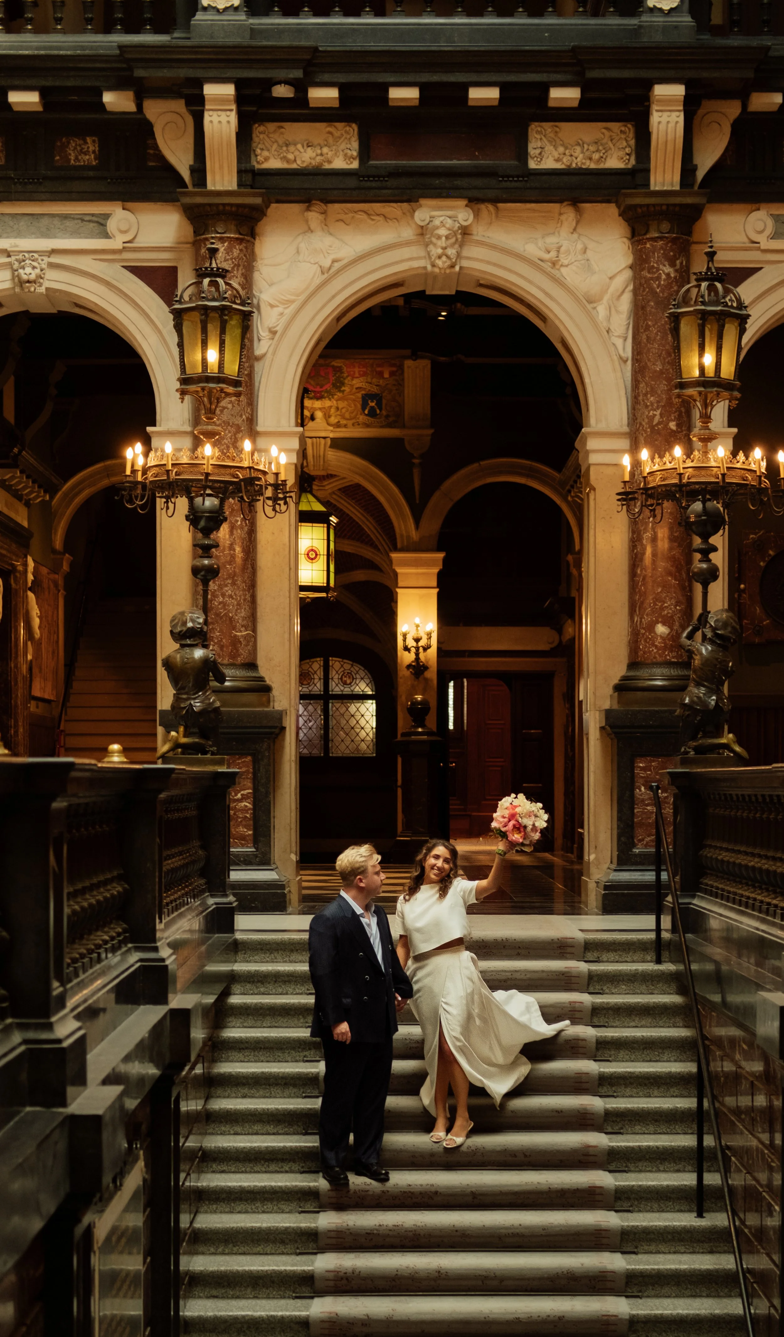 A woman in a wedding dress holding a bouquet of flowers standing on staircase with a man in a suit beside her inside a grand, historic building with ornate architecture and antique lighting fixtures.