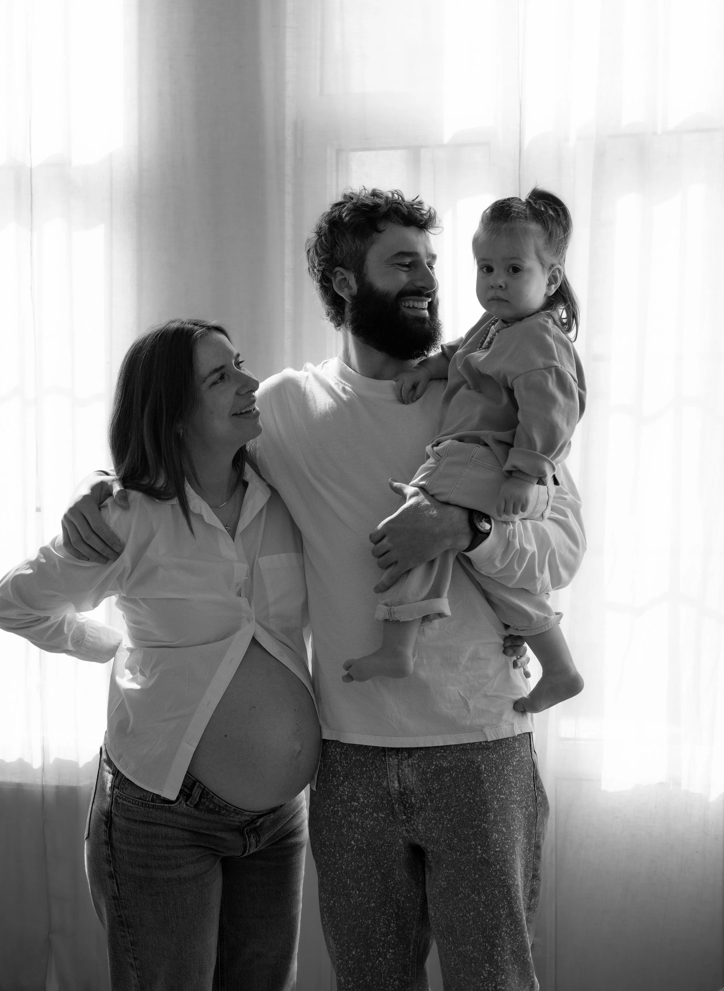 Black and white photo of a family of three standing near a window with sheer curtains. The pregnant woman on the left is smiling and looking at the man. The man in the center holds a young girl in his arms, who has a serious expression and is looking