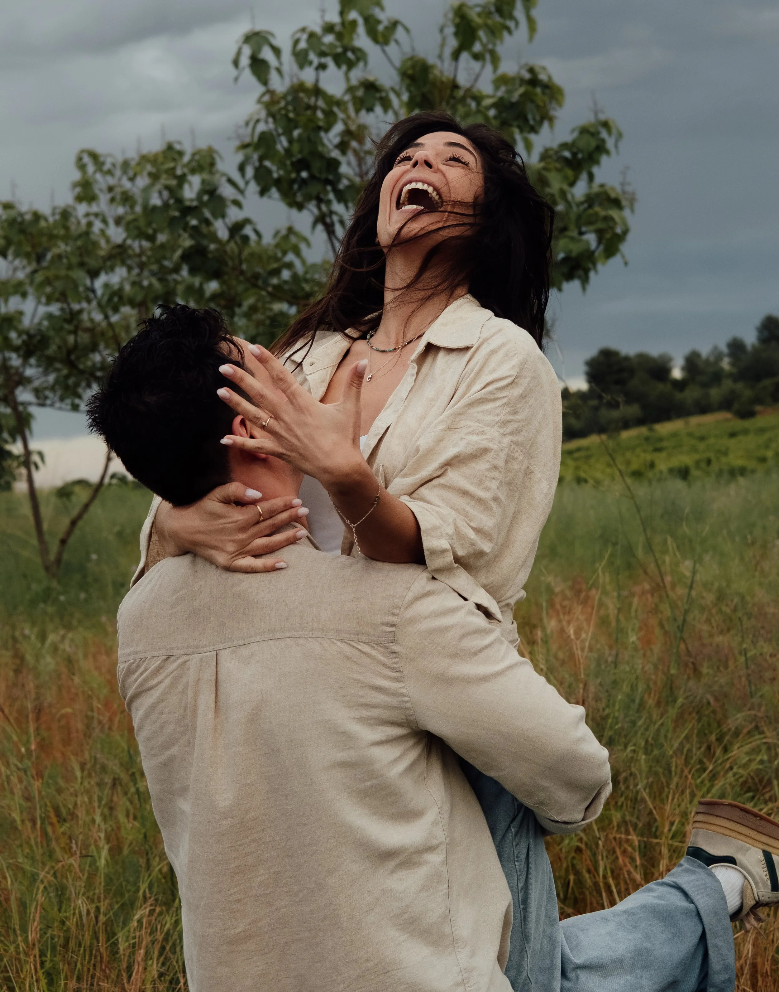 A woman laughing and embracing a person in the outdoors with trees and cloudy sky in the background.
