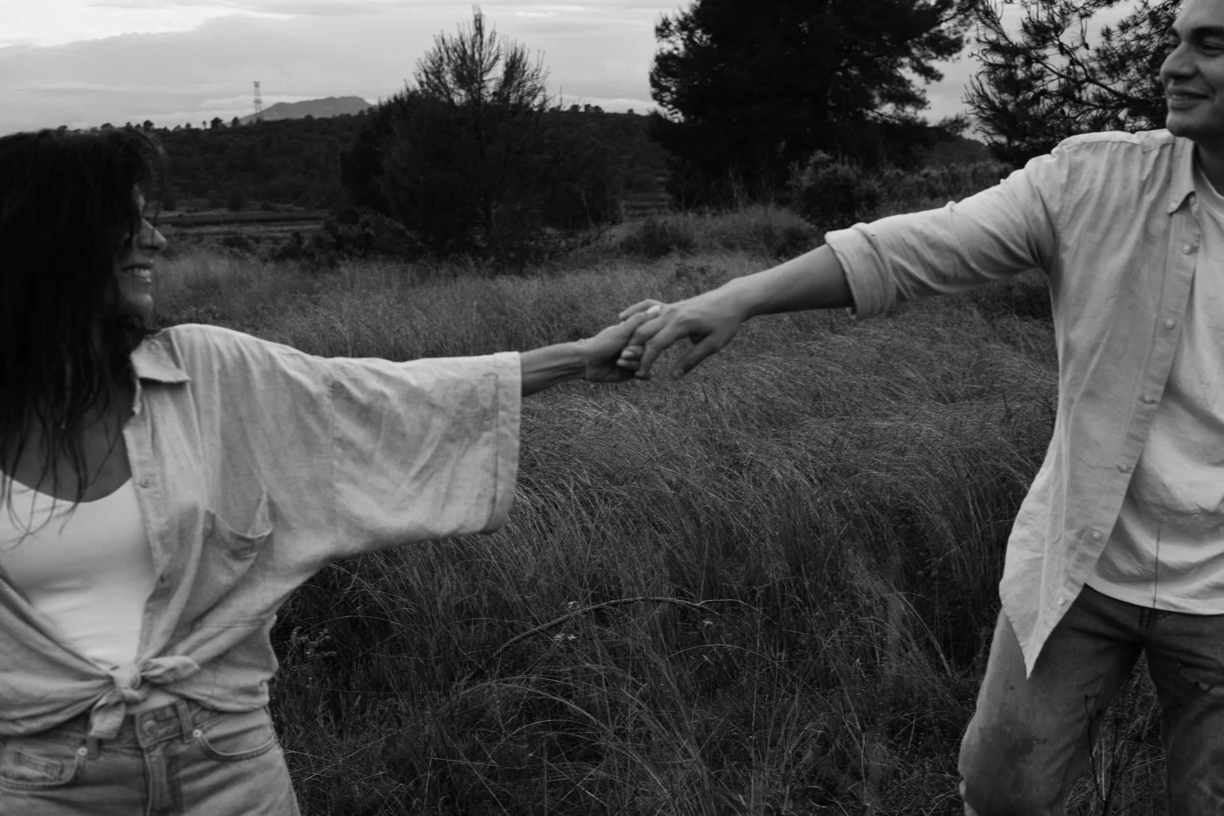 A black and white photo of a man and a woman holding hands in an outdoor field, smiling at each other. The woman has long hair and wears a tied shirt, while the man wears a button-up shirt. Trees and hills are visible in the background.