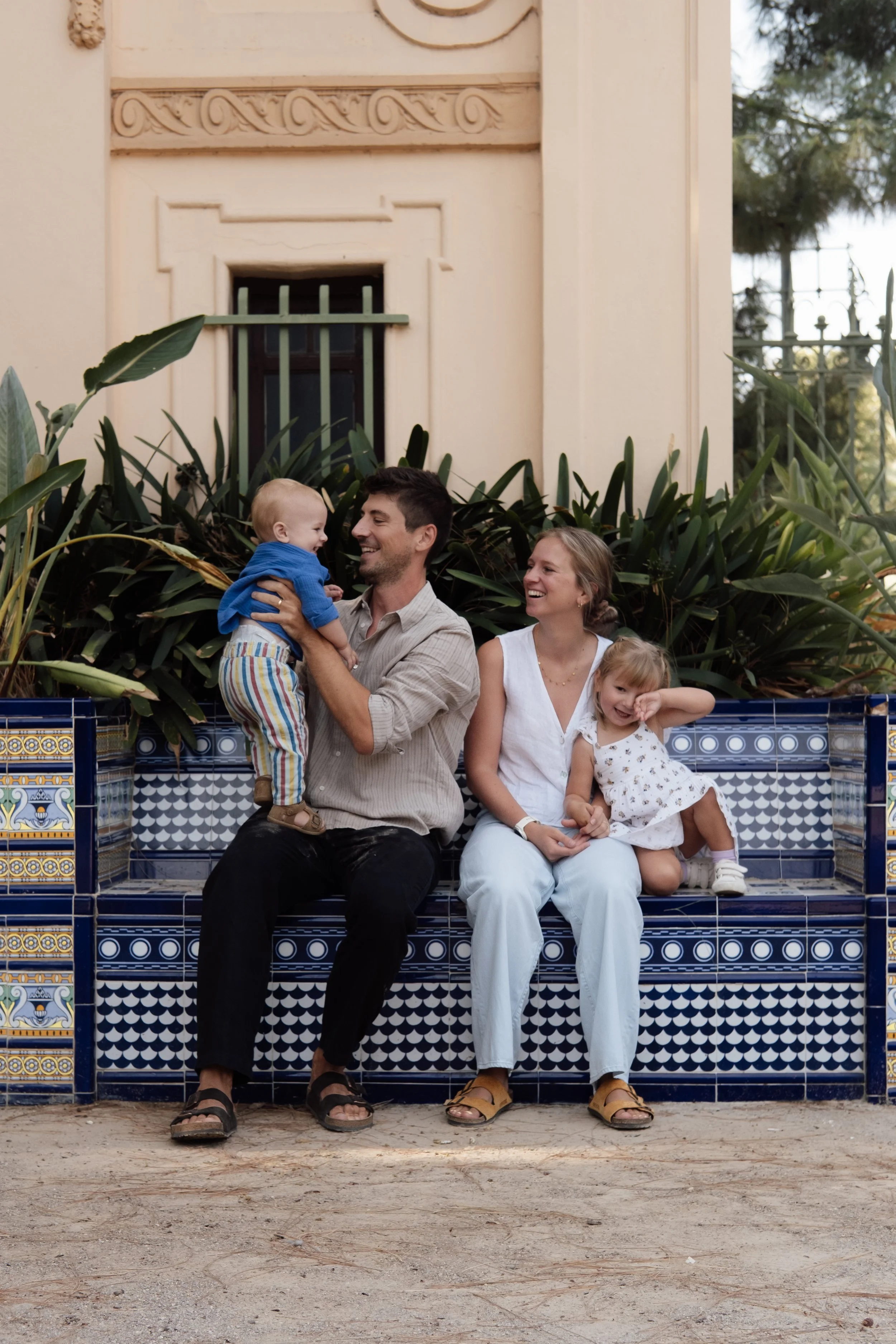A man holding a young boy in a blue sweater and striped pants, smiling at each other, with a woman and a young girl sitting on patterned blue and white tiled steps behind them, smiling and enjoying their time together. There are green plants and a beige building with decorative architectural details in the background.