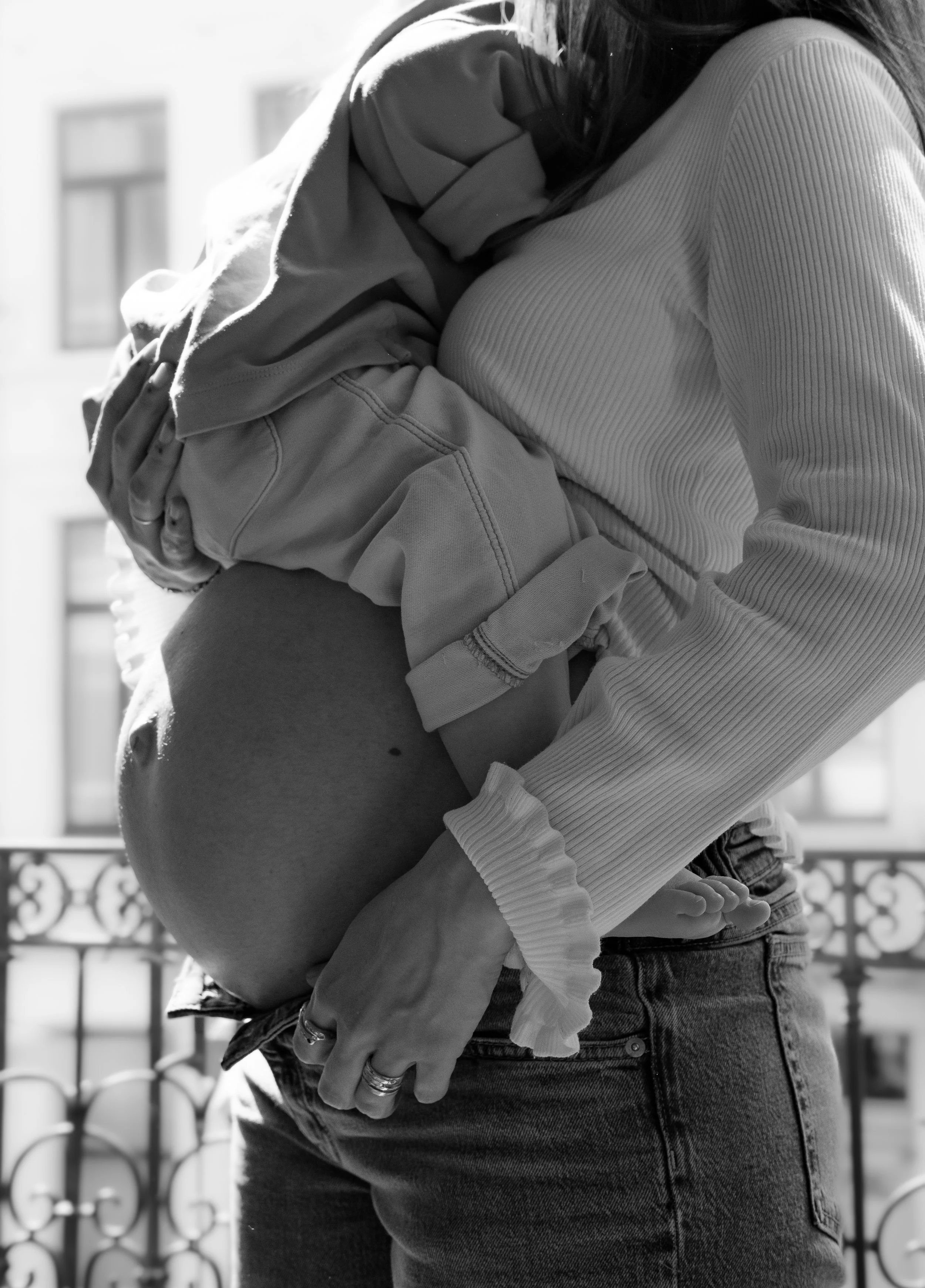 A woman with long hair wearing a striped long-sleeve shirt and jeans hugging a pregnant woman with a hoodie and arm tattoos, standing outside with a decorative metal railing in the background.