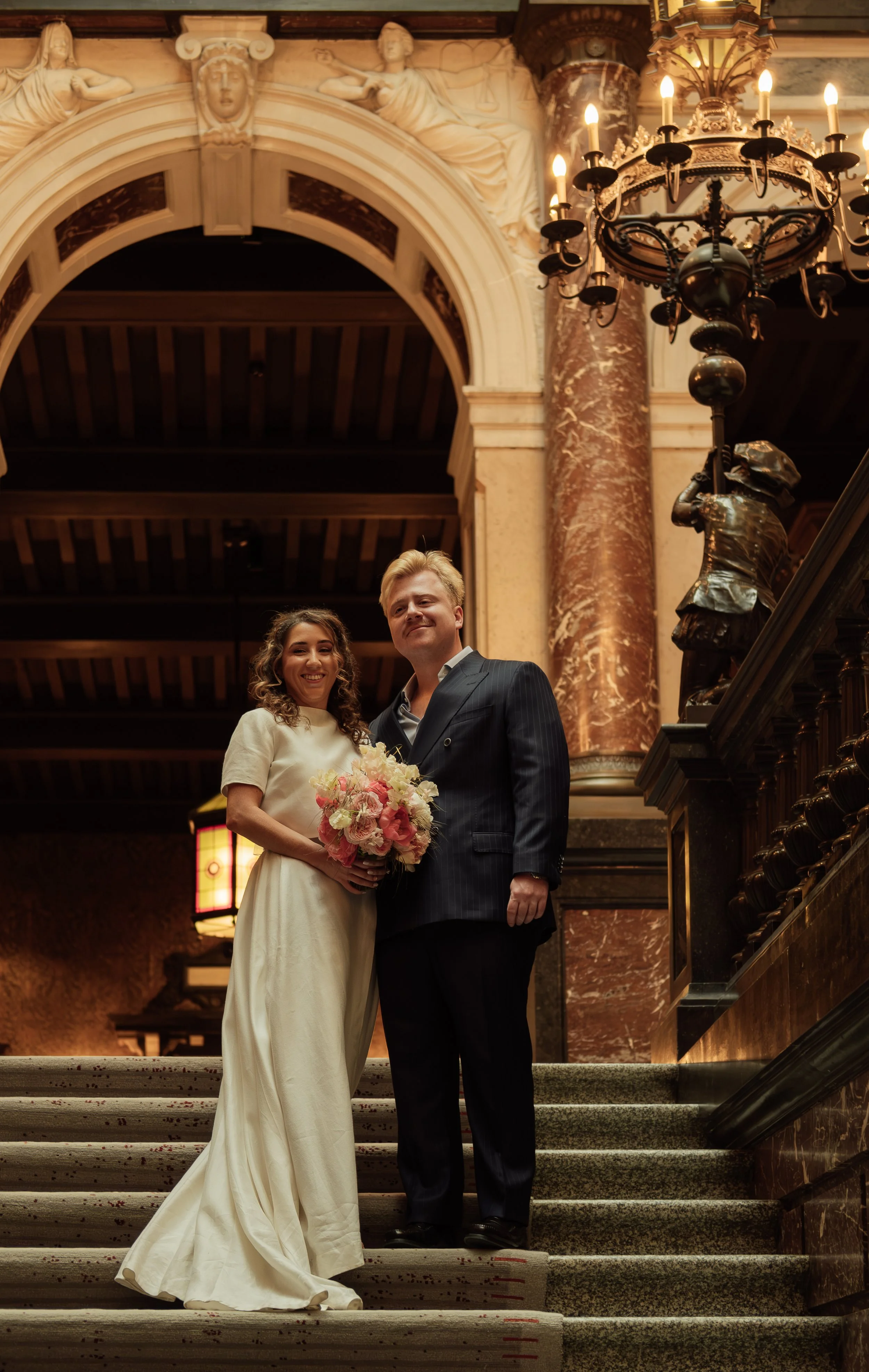 A newlywed couple standing on a staircase in a grand, ornate building. The bride is holding a bouquet of pink and white flowers and wearing a white wedding dress. The groom is dressed in a dark pinstripe suit. The background features elaborate archit