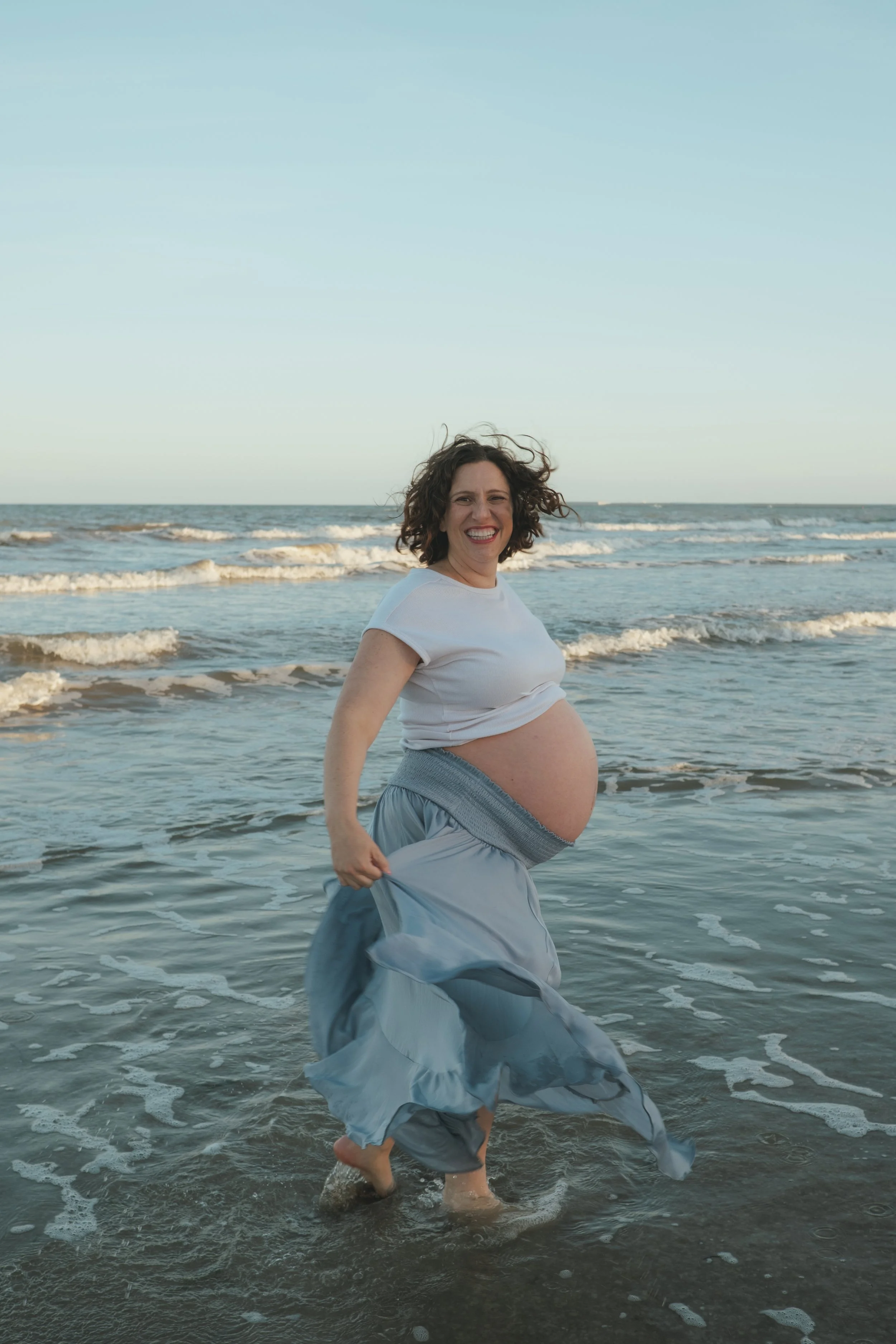 Smiling pregnant woman in a flowing dress standing in ocean waves at the beach