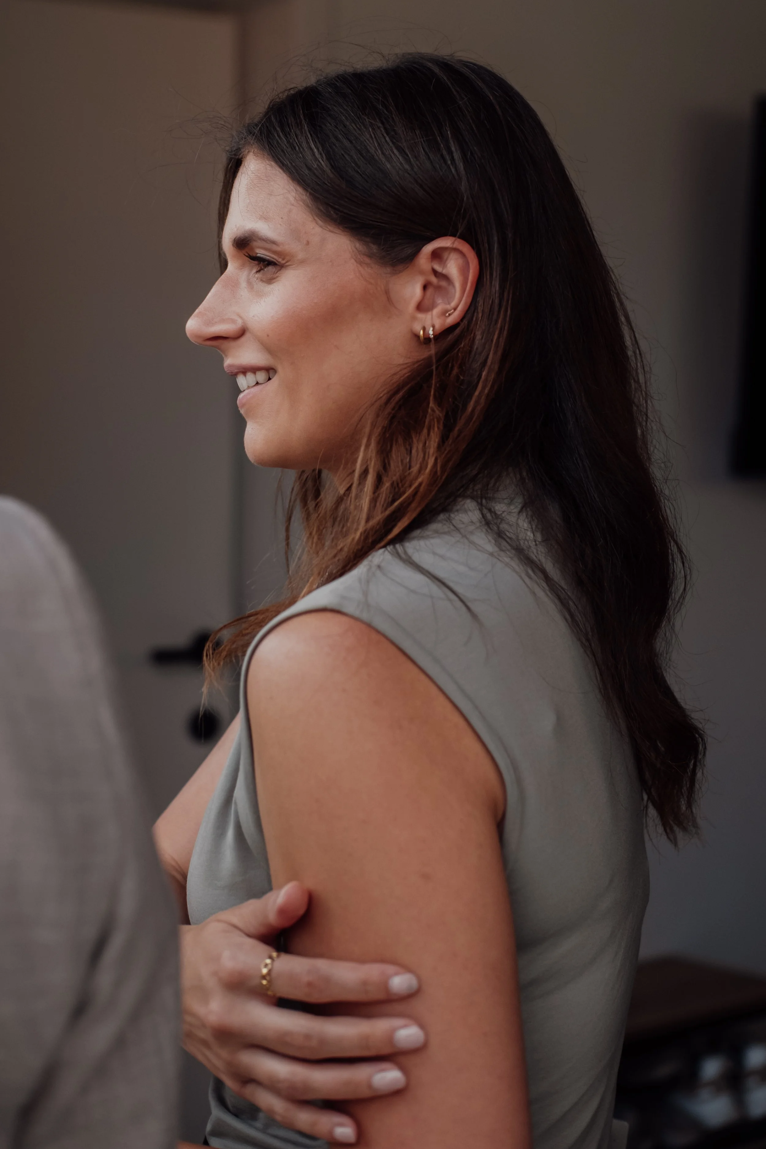 A woman with dark brown hair smiling and facing left, wearing earrings and a light gray sleeveless top, touching her upper arm.