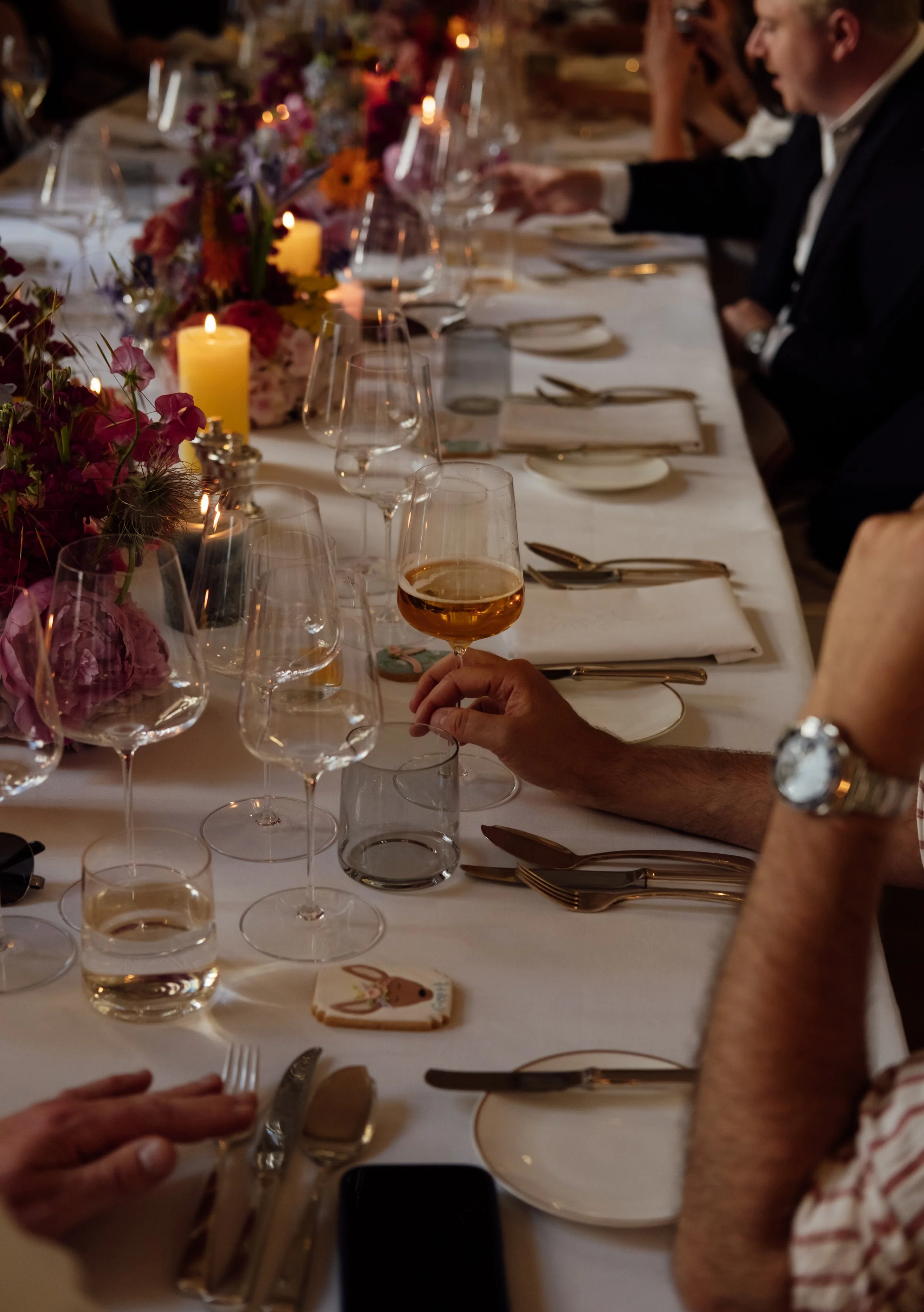 A formal dinner setting on a long table decorated with flowers, candles, and glassware, with people dressed in formal attire enjoying a meal.