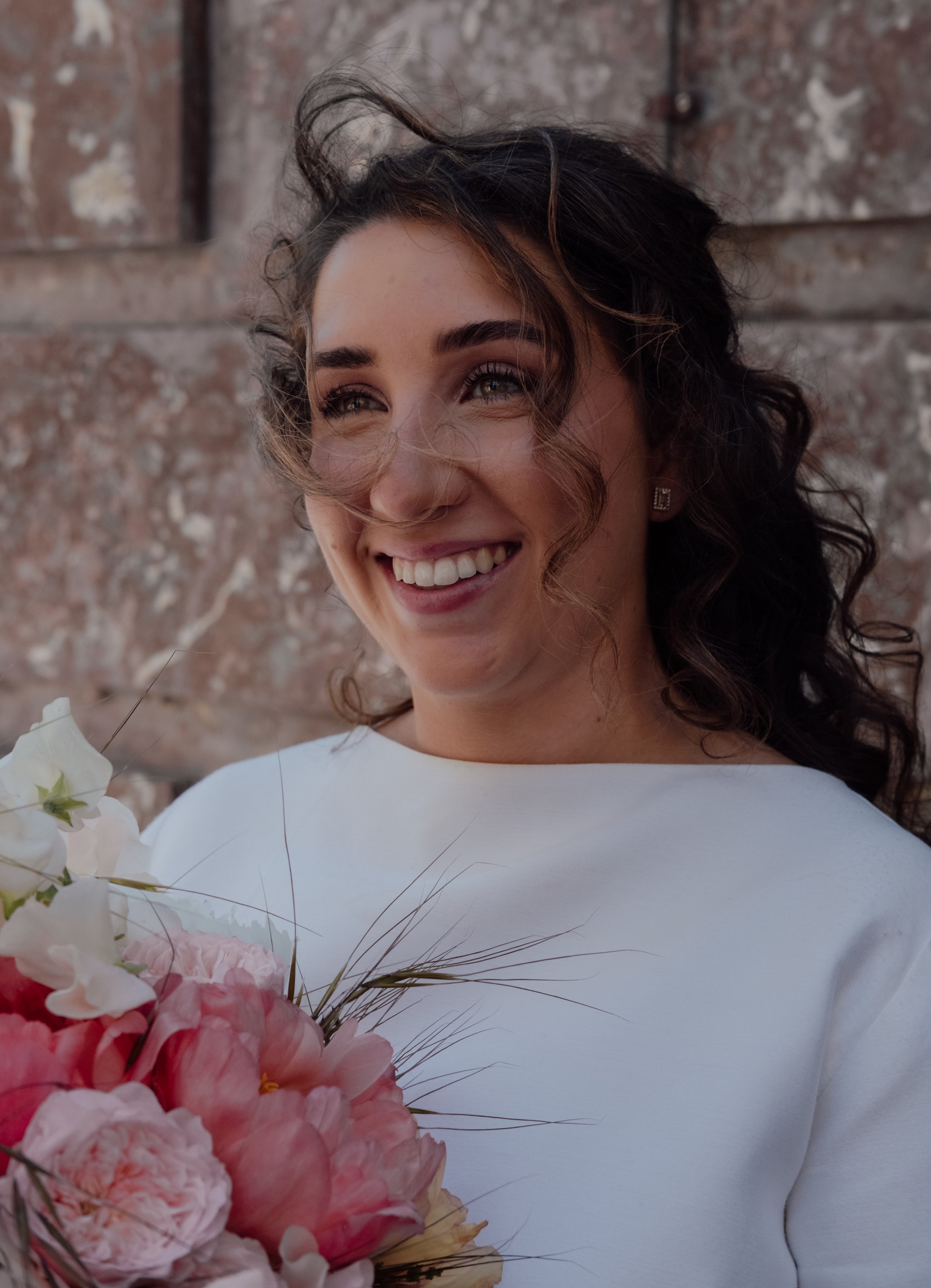Close-up of a smiling woman with dark curly hair and earrings, holding flowers against a brick wall background.