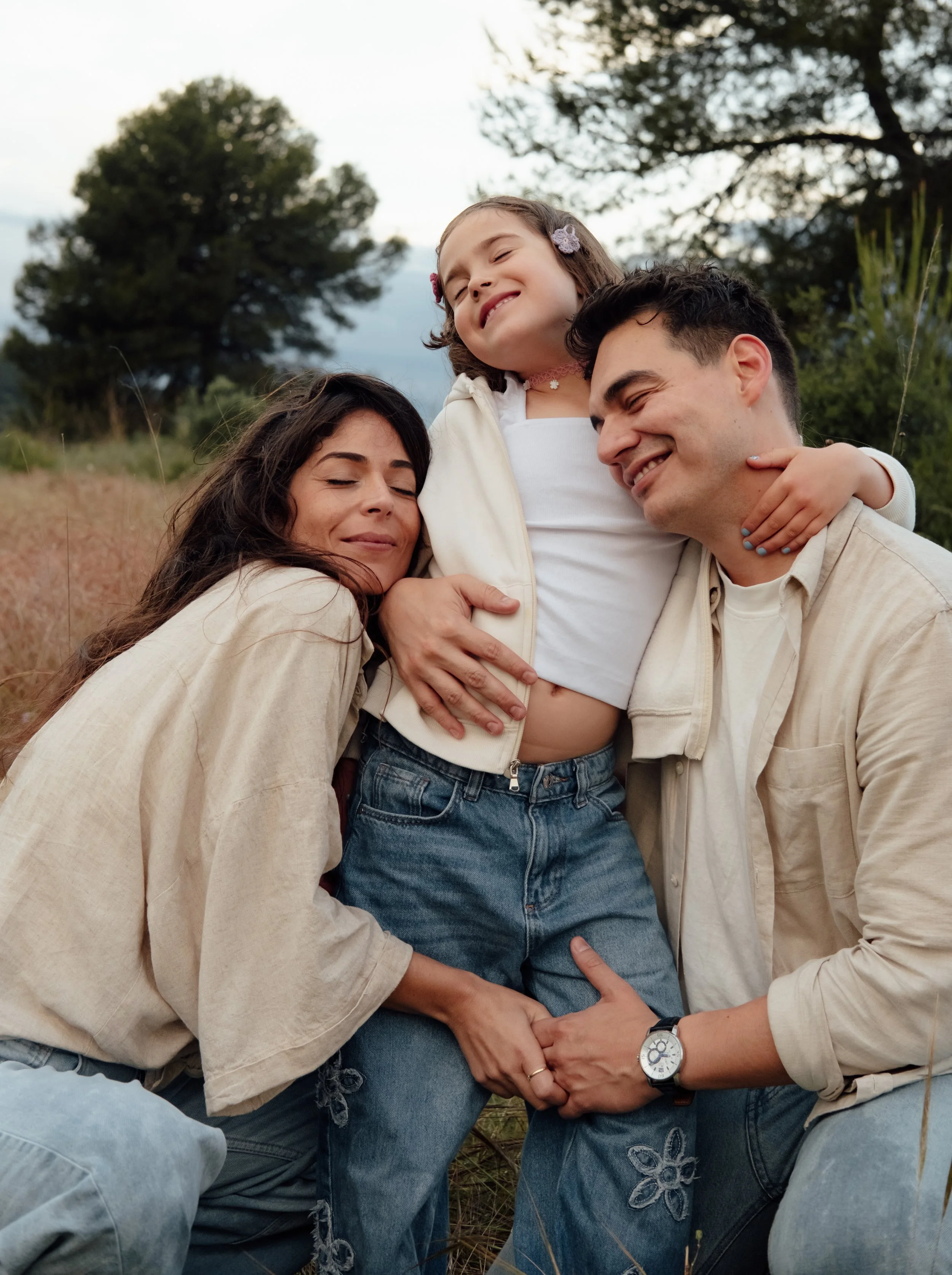 A family of three, a woman, a man, and a young girl, enjoying a moment outdoors in a grassy area with trees. The woman and man are smiling, and the girl is happy with her eyes closed, hugging the man.