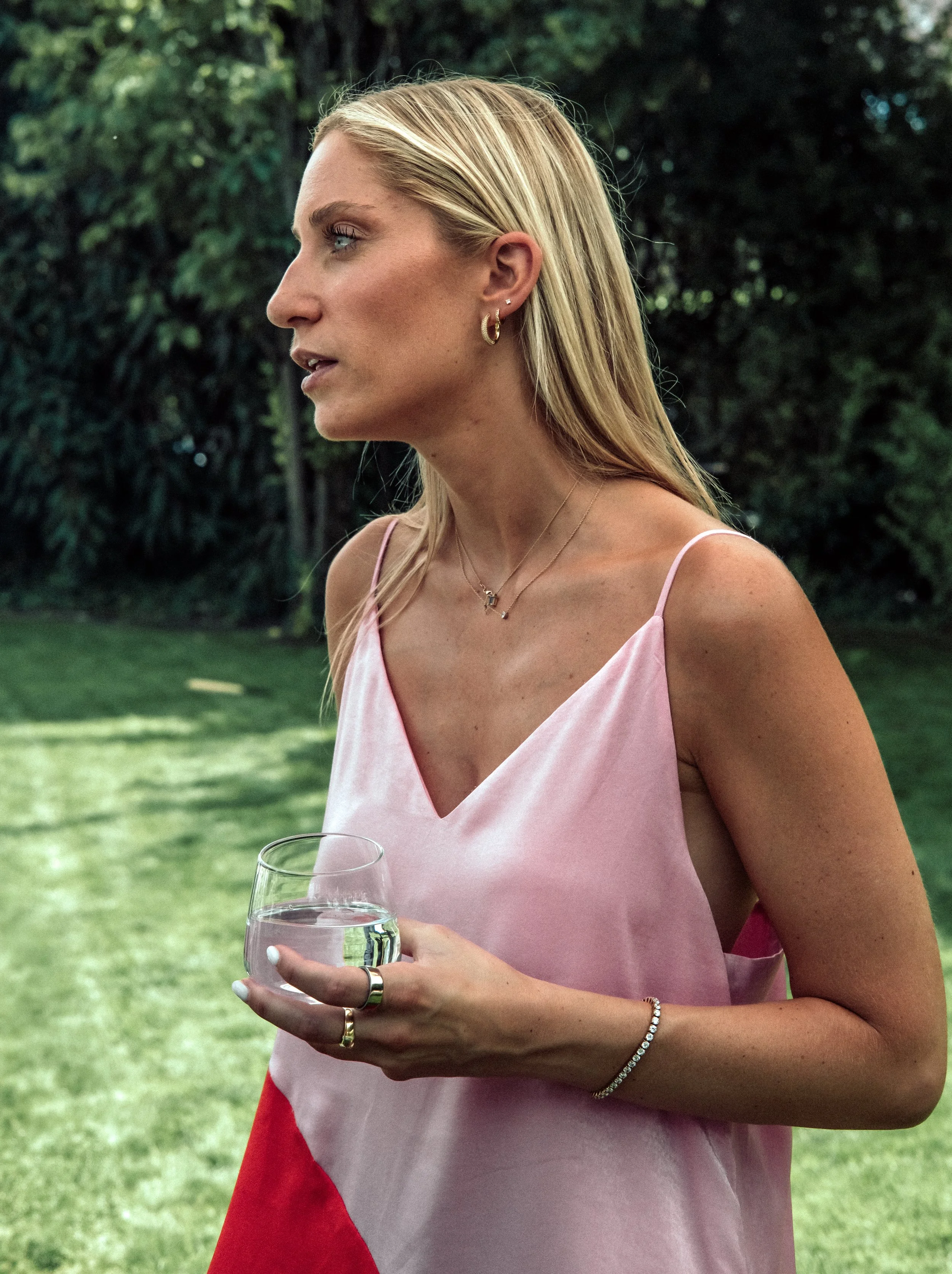 Side profile of a blonde woman wearing pink dress, holding a glass of water, outdoors with green trees in the background.
