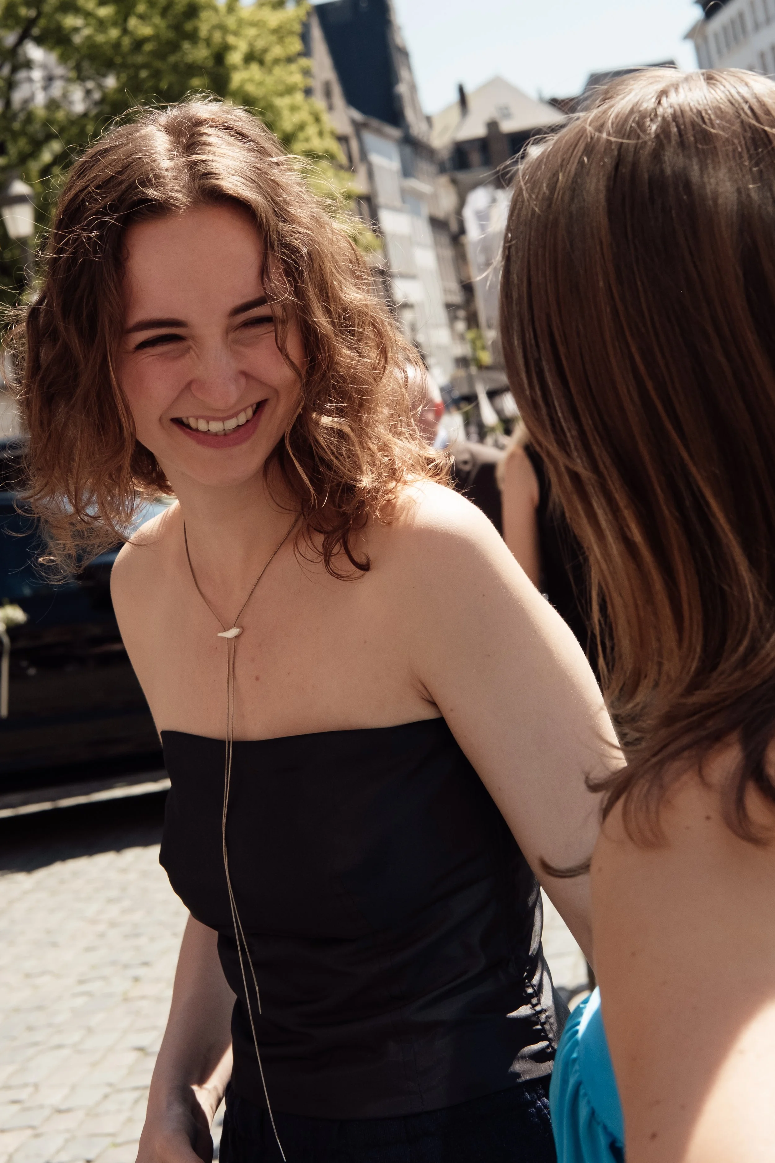 Two women smiling and talking outdoors on a sunny day, with one in a black strapless top and the other in a blue sleeveless dress.