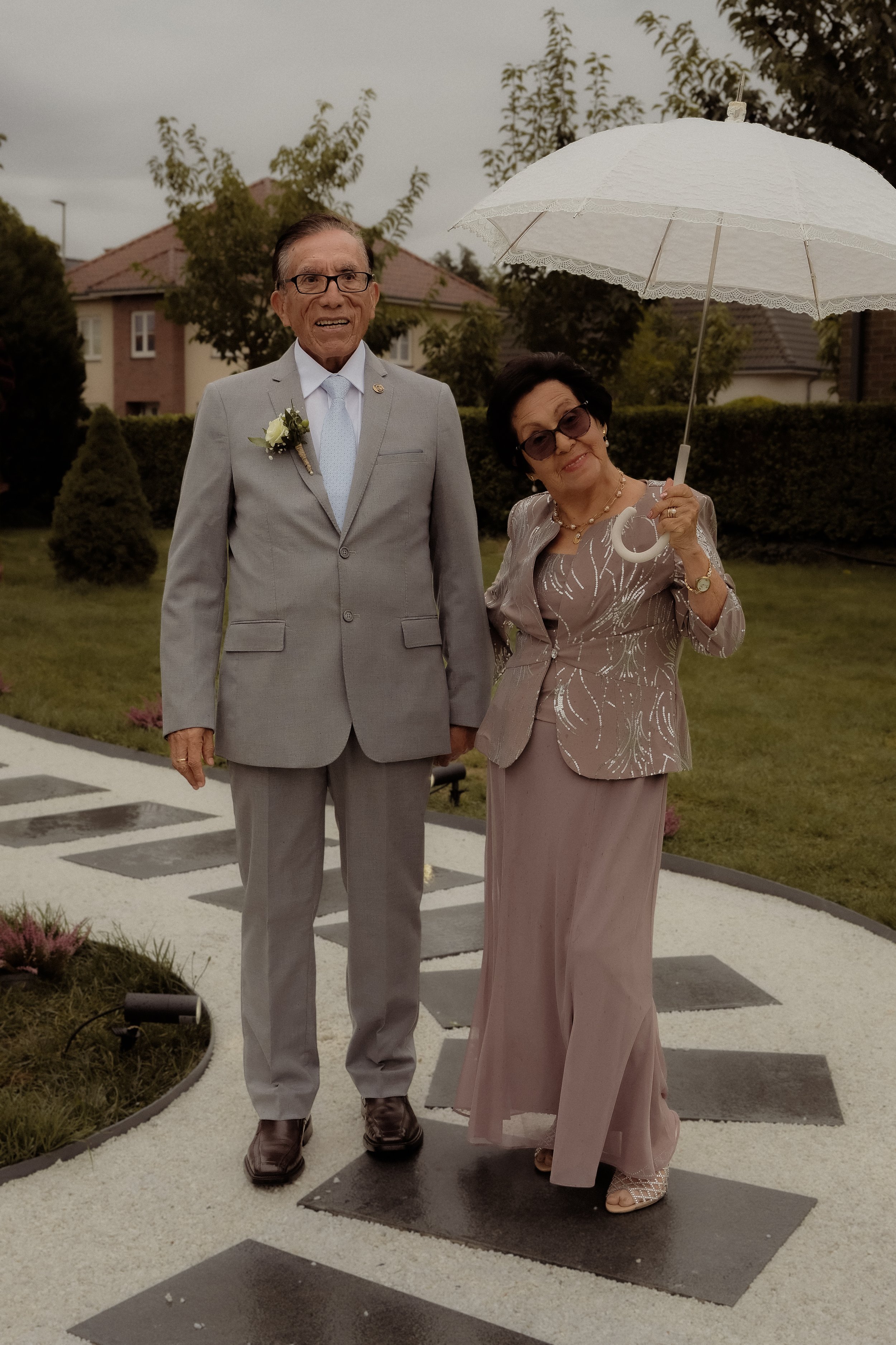 An elderly man in a light gray suit with a boutonniere and an elderly woman in a pink dress and jacket with a white umbrella standing on a decorated outdoor walkway.