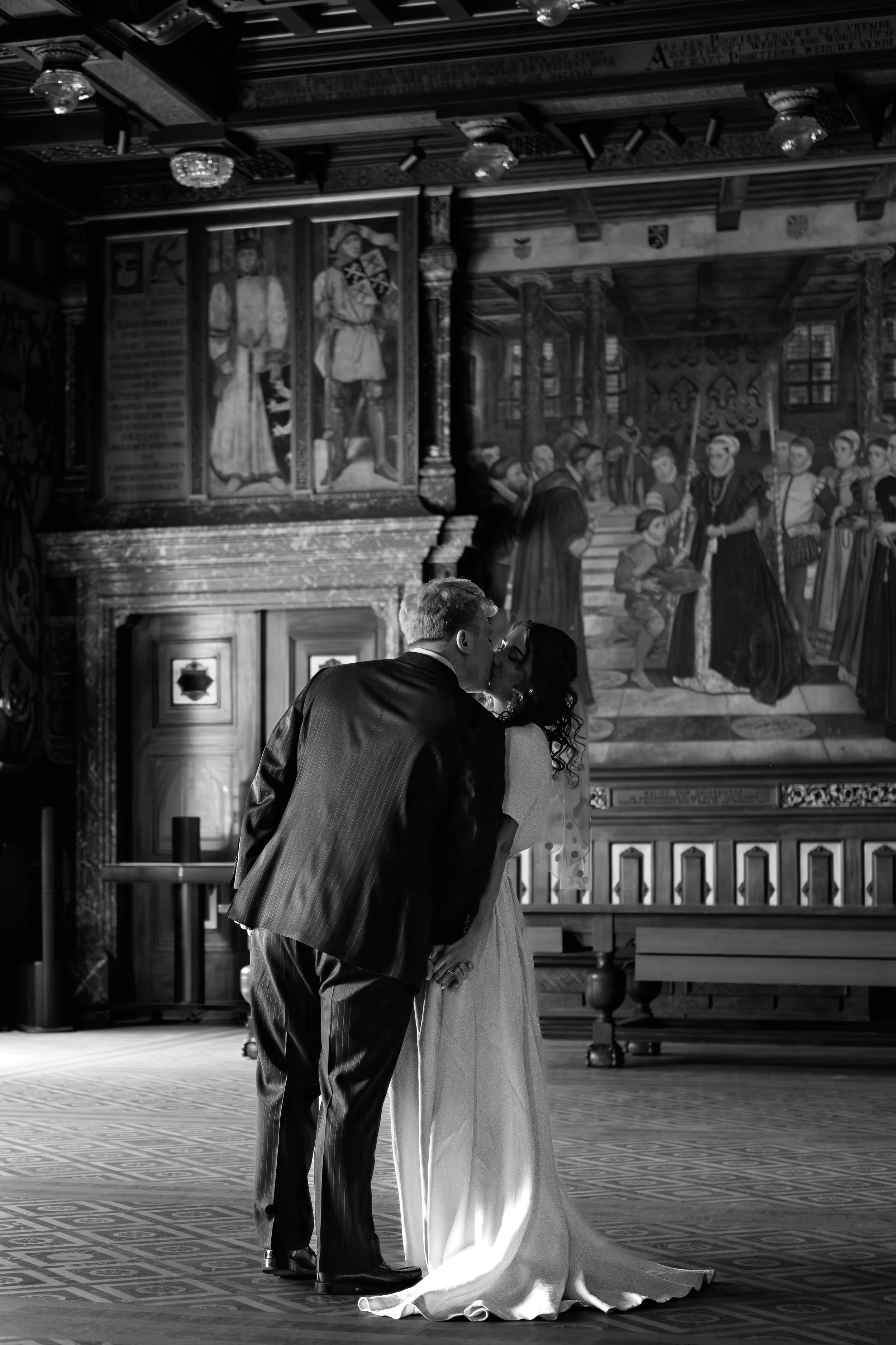 A couple in wedding attire sharing a kiss in a decorated hall with a large mural of historical figures on the wall behind them.