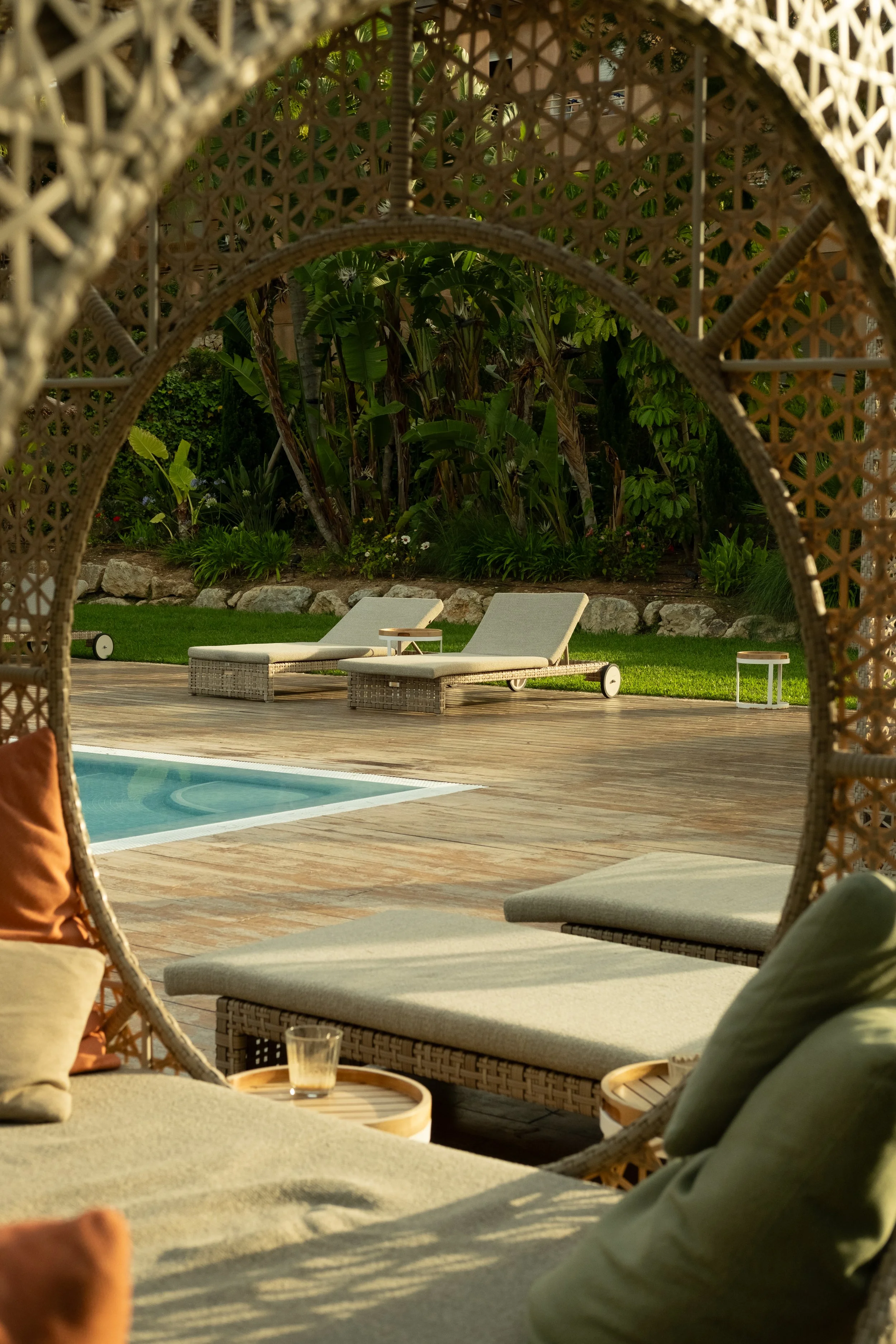 A backyard scene viewed through a wicker and rattan arch with seating cushions, a table, and a small tray with a glass. In the background, there are lounge chairs, a pool, grass, and lush green tropical plants and trees.