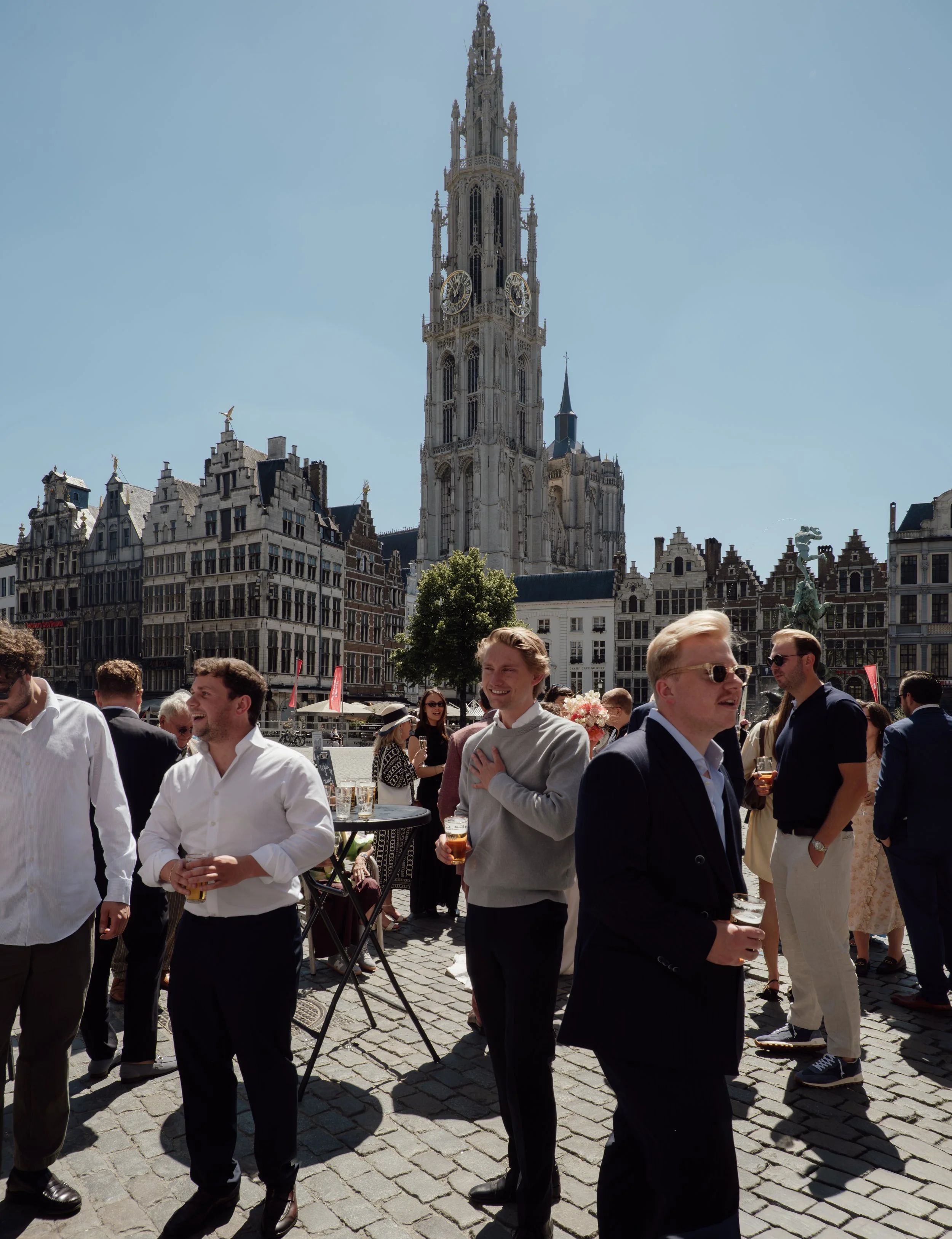 People gathered outdoors in a city square with historical buildings and a tall clock tower in the background. The sky is clear and sunny.
