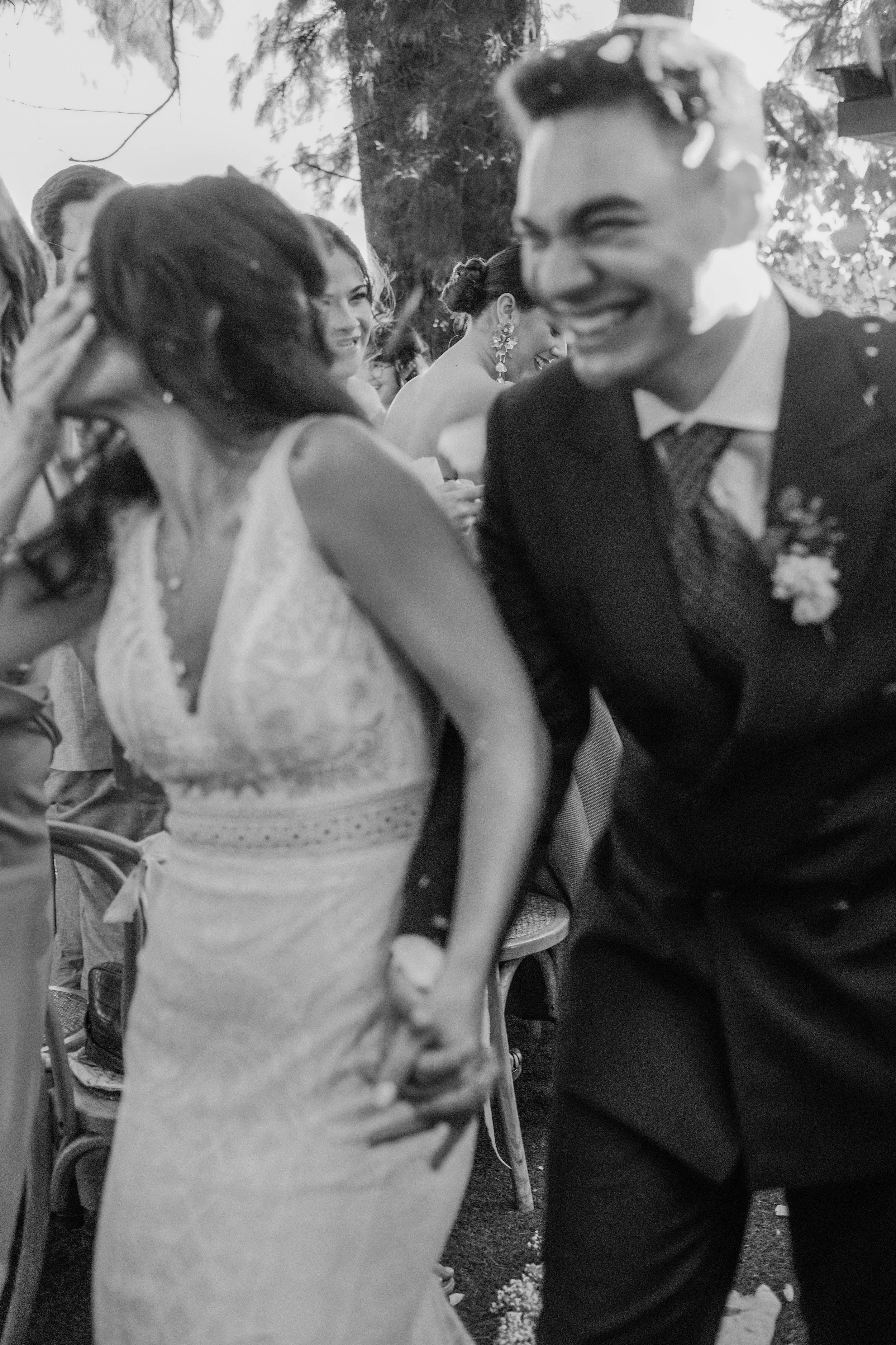 Black and white photo of a group of people at a social gathering outdoors. A man in a suit and tie is smiling and appears to be in conversation. Several women are nearby, smiling and engaging with each other. The background features trees and outdoor setting.