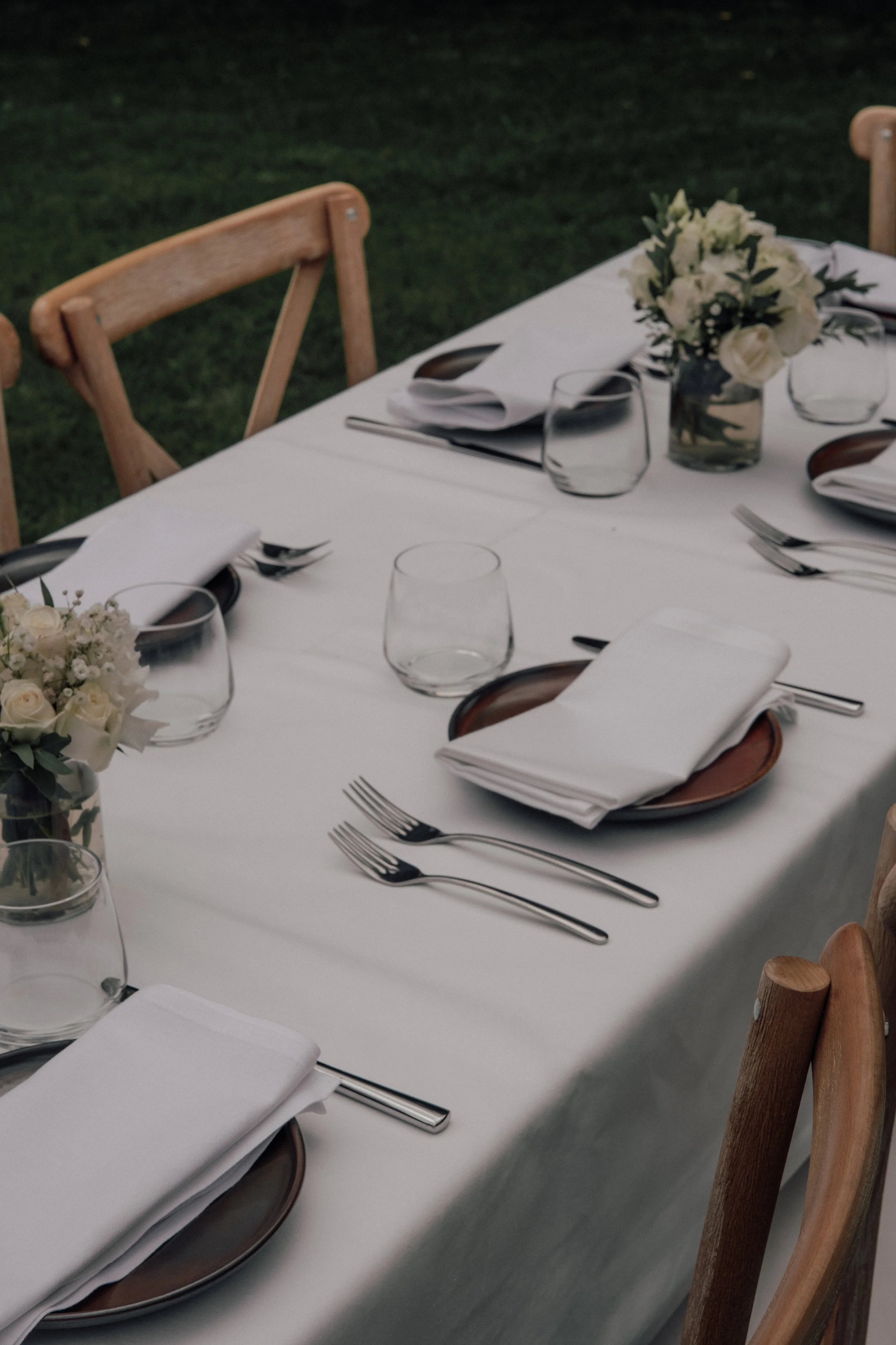 A formal dining table set outdoors on grass with white tablecloth, floral centerpiece, white napkins, and silverware, surrounded by wooden chairs.