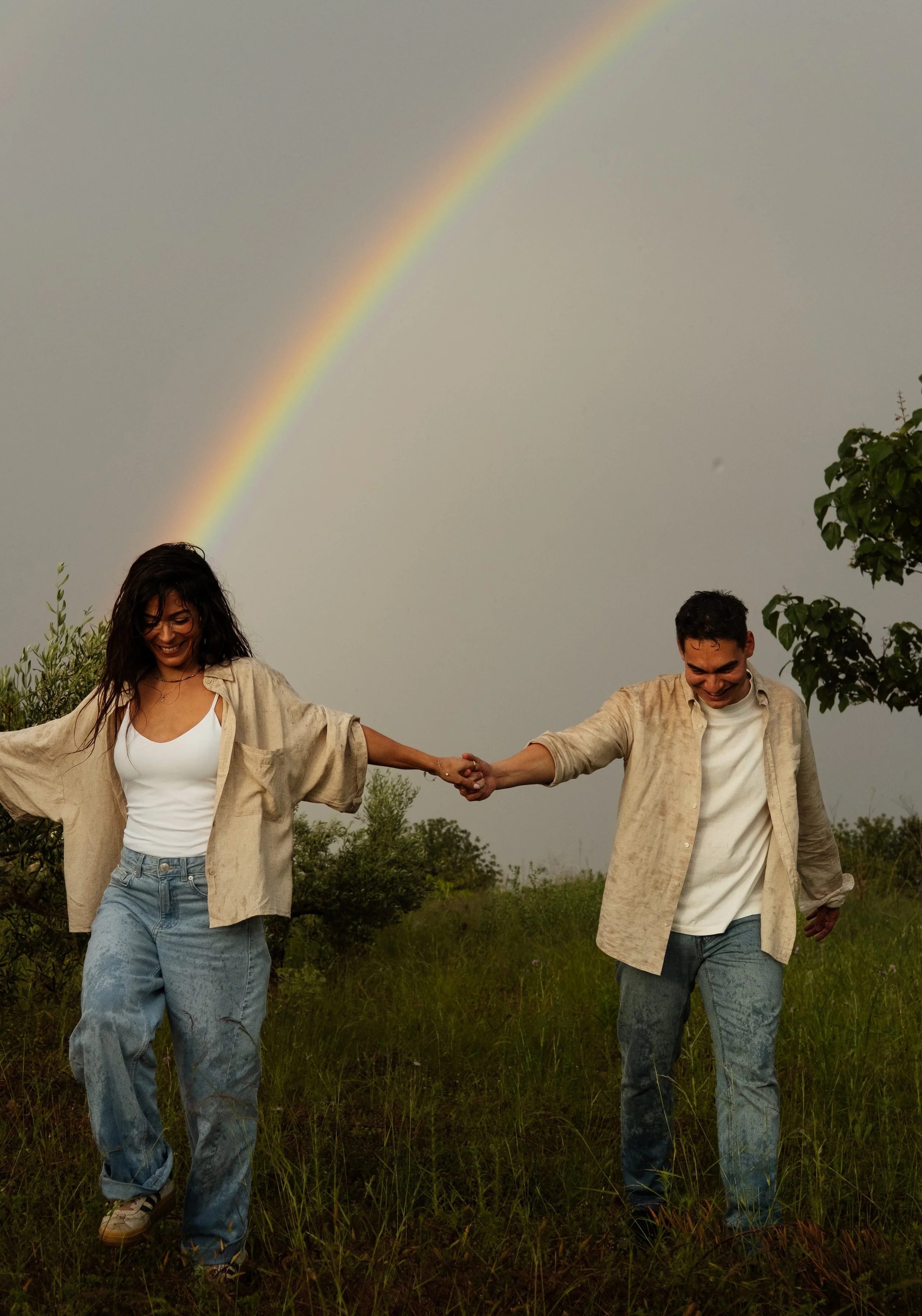 A smiling woman and man holding hands and walking through a grassy field, with a rainbow in the cloudy sky above.