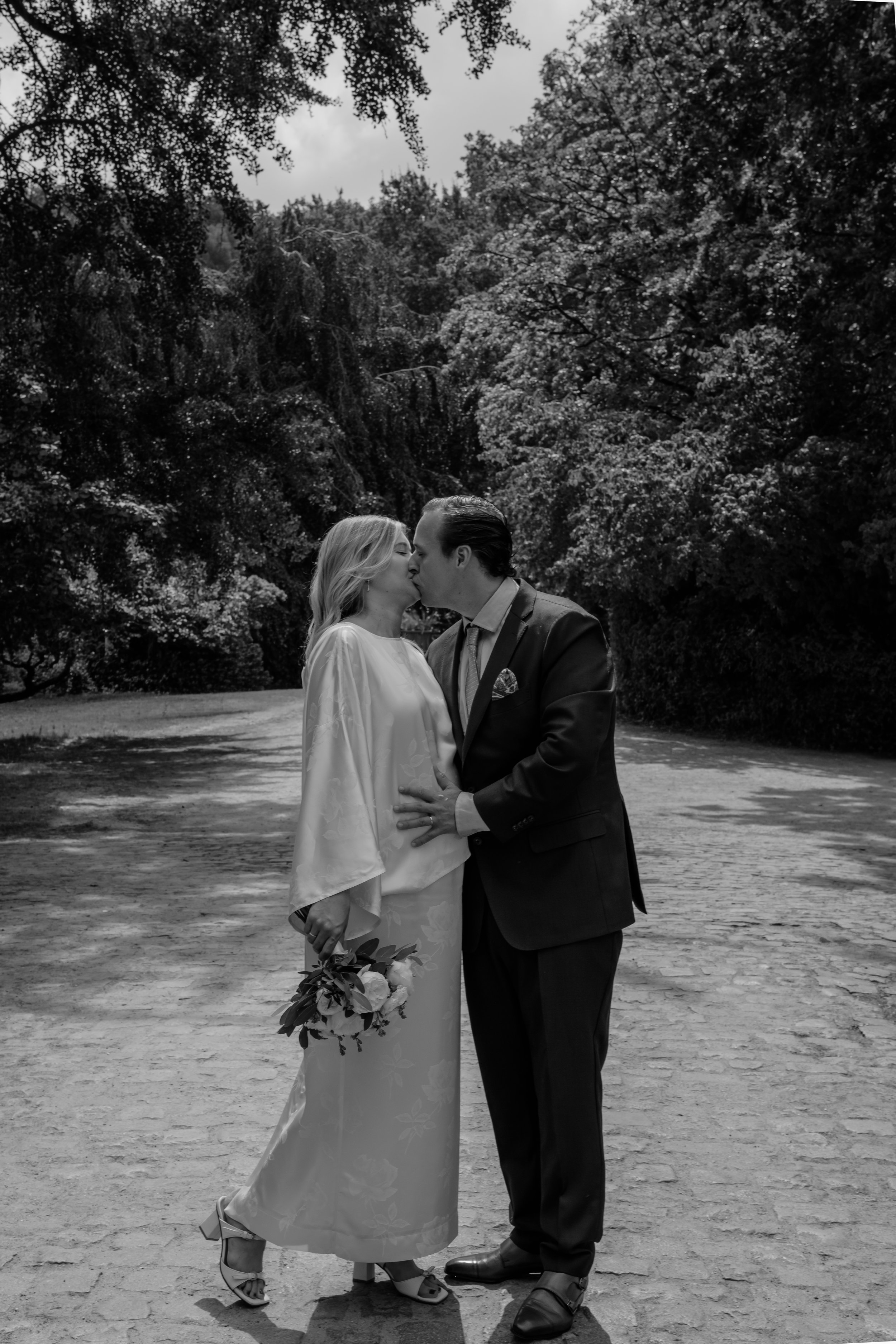 A black and white photograph of a couple kissing outdoors, with trees in the background. The woman is wearing a long dress and holding a bouquet, and the man is wearing a suit.