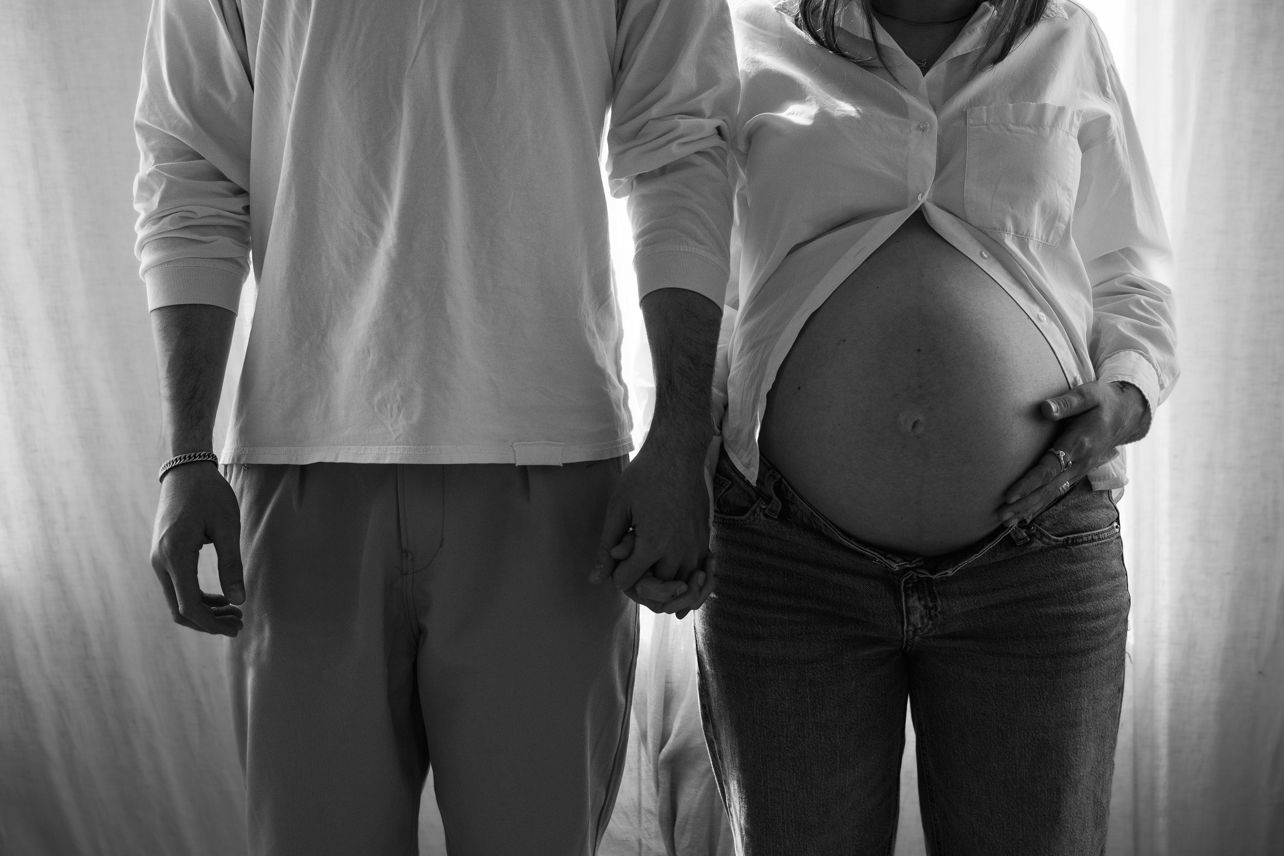 A pregnant woman and a man holding hands, standing side by side in front of a light-colored background.