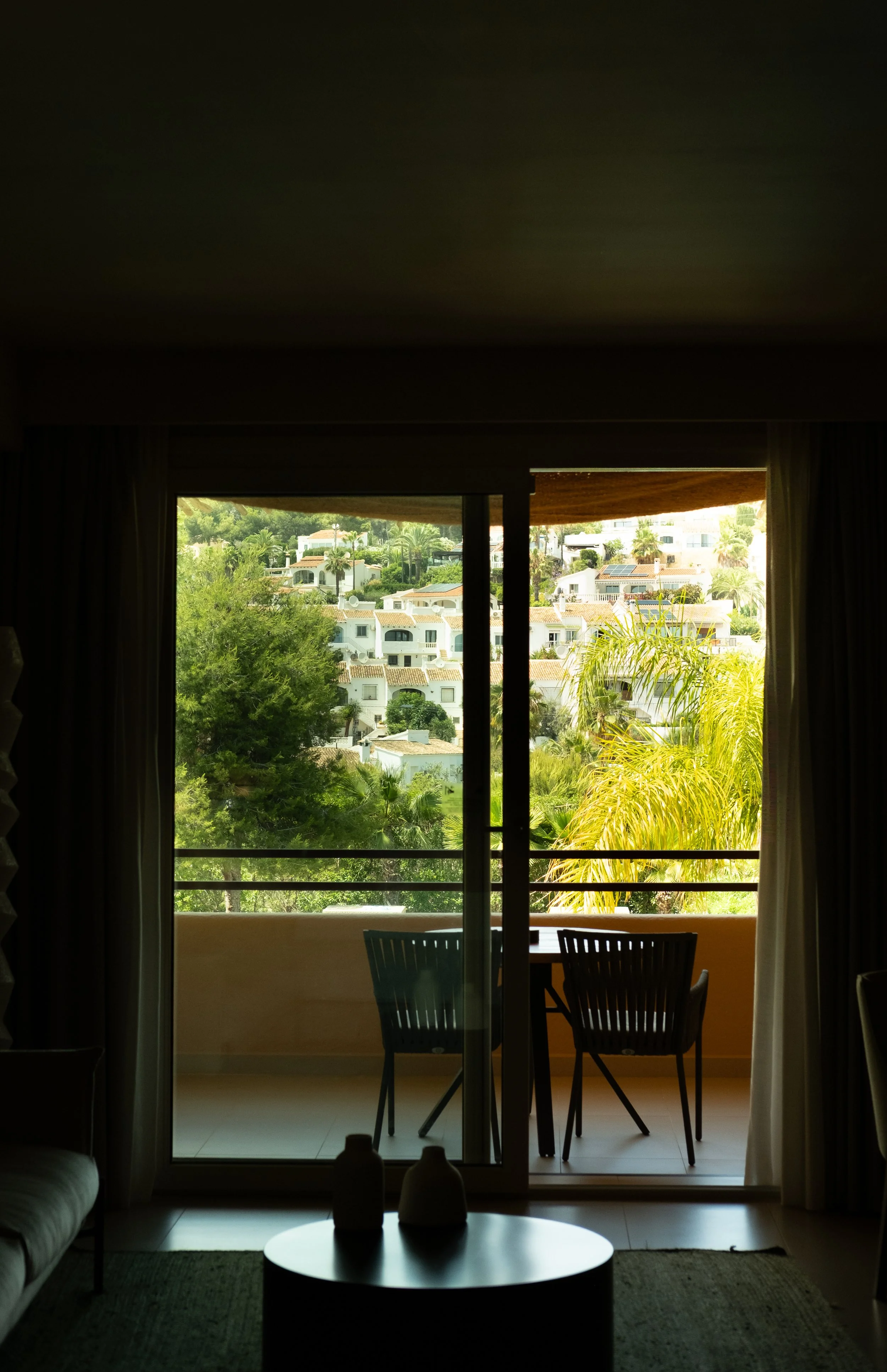 Interior view of a living room with a glass sliding door leading to a balcony, overlooking a hillside neighborhood with white buildings, green trees, and some palm trees.