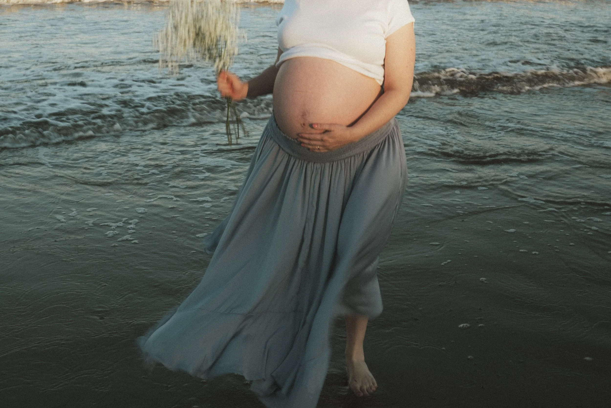 Pregnant woman in a white top and flowing gray skirt standing in shallow ocean water
