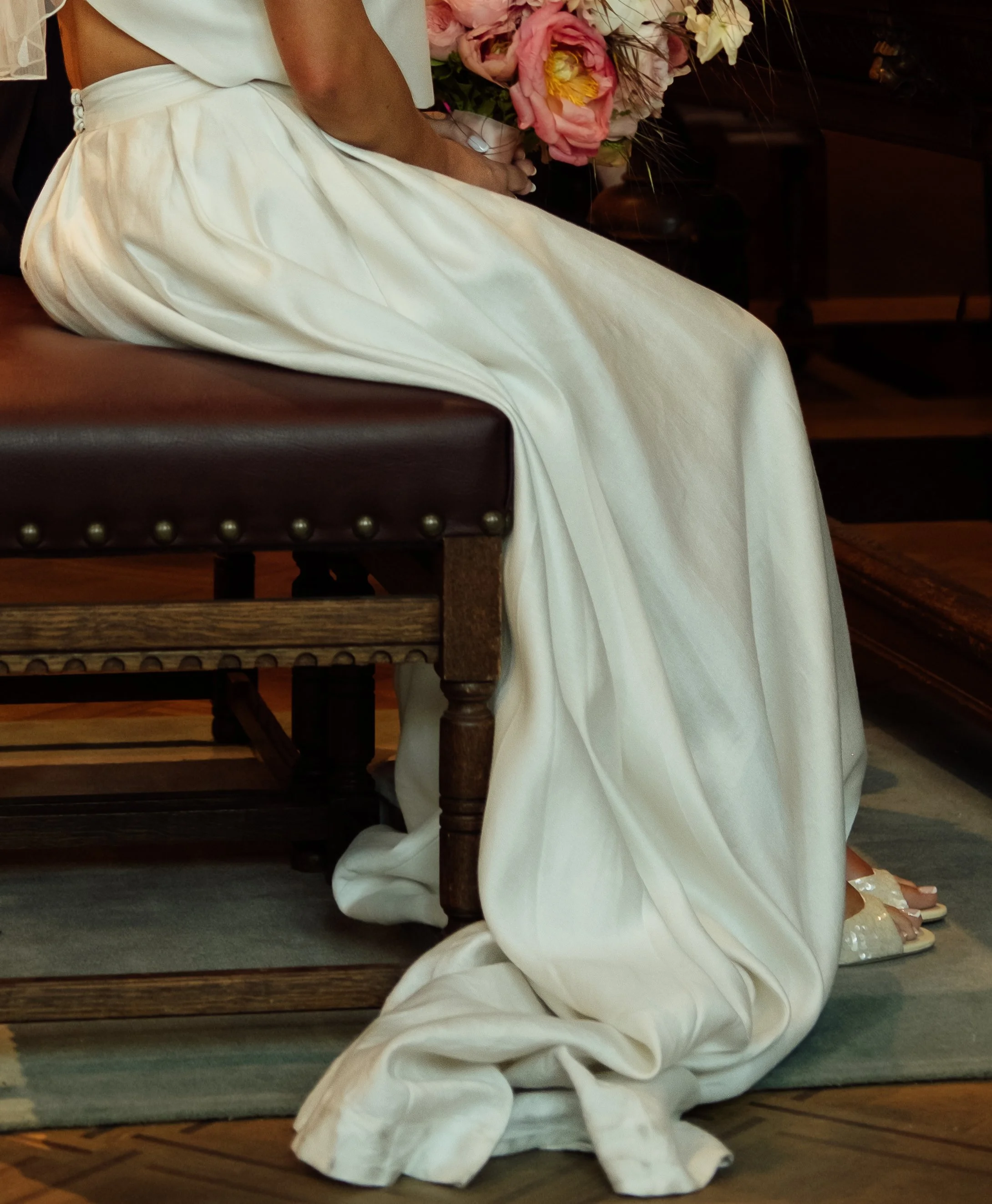 A woman kneeling on a rug, wearing a long, white dress, next to a framed flower arrangement with pink and white flowers, and a small row of white shoes on the floor.